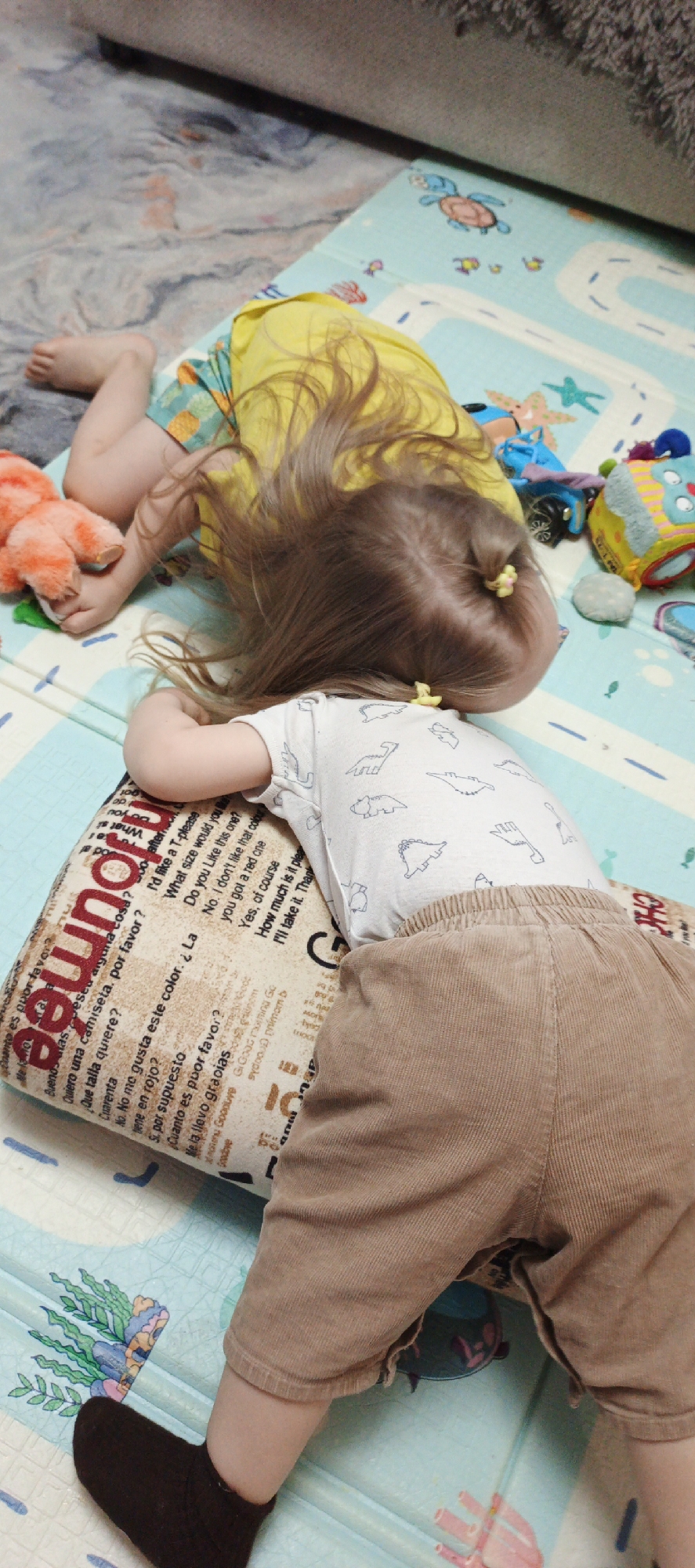 Two young children are sleeping on a light blue and white patterned play mat. One child is wearing a yellow shirt and patterned bottoms, while the other is wearing a white shirt with dinosaur outlines and brown corduroy shorts, with one black sock visible on their left foot. This child is resting their head on a patterned pillow with text in different languages. There are several toys scattered around the children on the play mat. The location is presumed to be indoors, likely a home setting. There is no evidence to indicate the city of Chaikovsky, Russia.