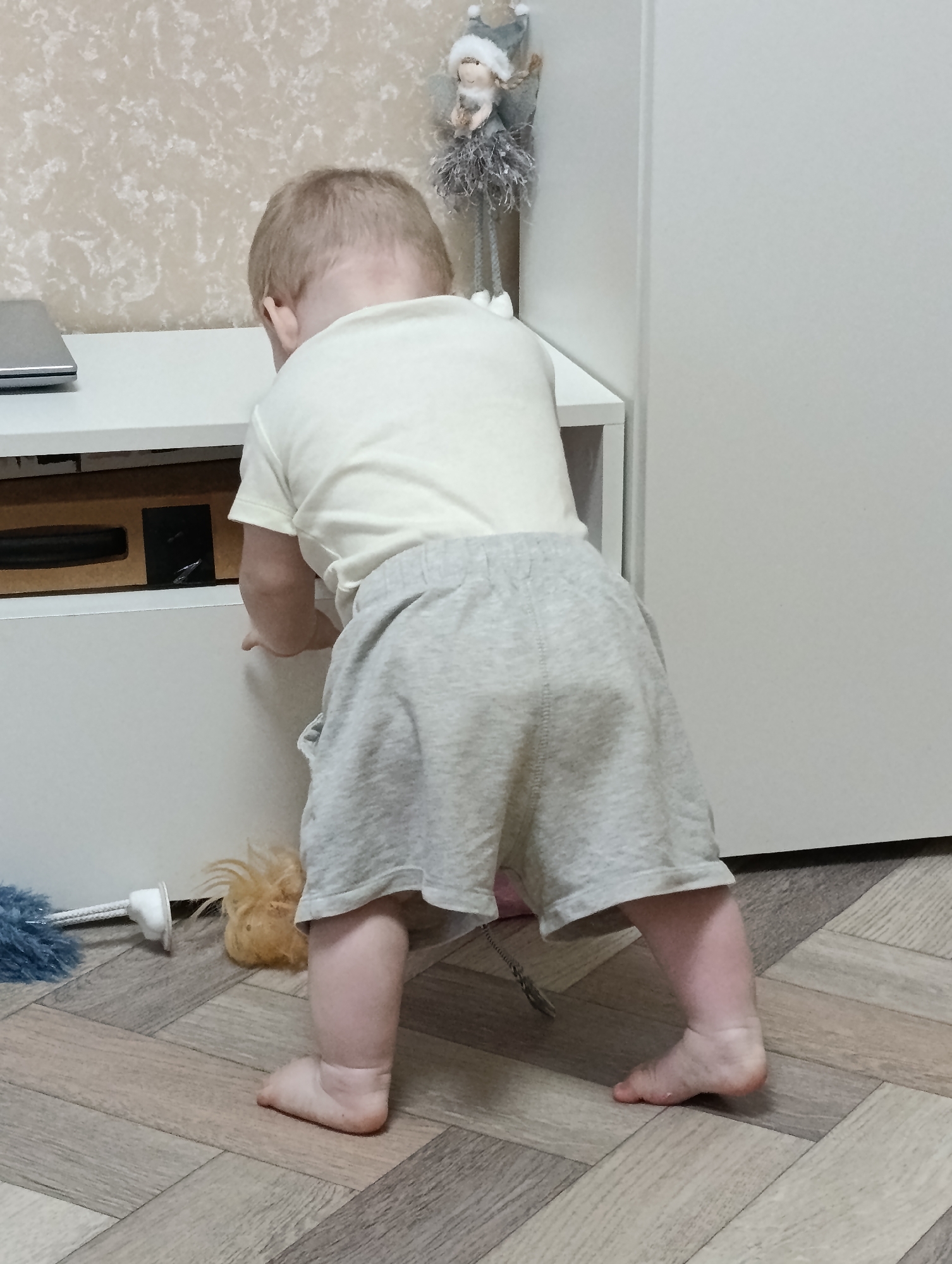 A young child, viewed from the rear, is standing and reaching towards a white cabinet. The child has blonde hair and is wearing a light-colored t-shirt and light gray shorts. The child's feet are bare. To the left of the child, on the wooden floor, are a toy brush with blue bristles and a fluffy orange toy. The background consists of a wall with a textured pattern and a white cabinet. The scene appears to be indoors. This image does not contain enough information to determine its location in Chaikovsky, Russia.