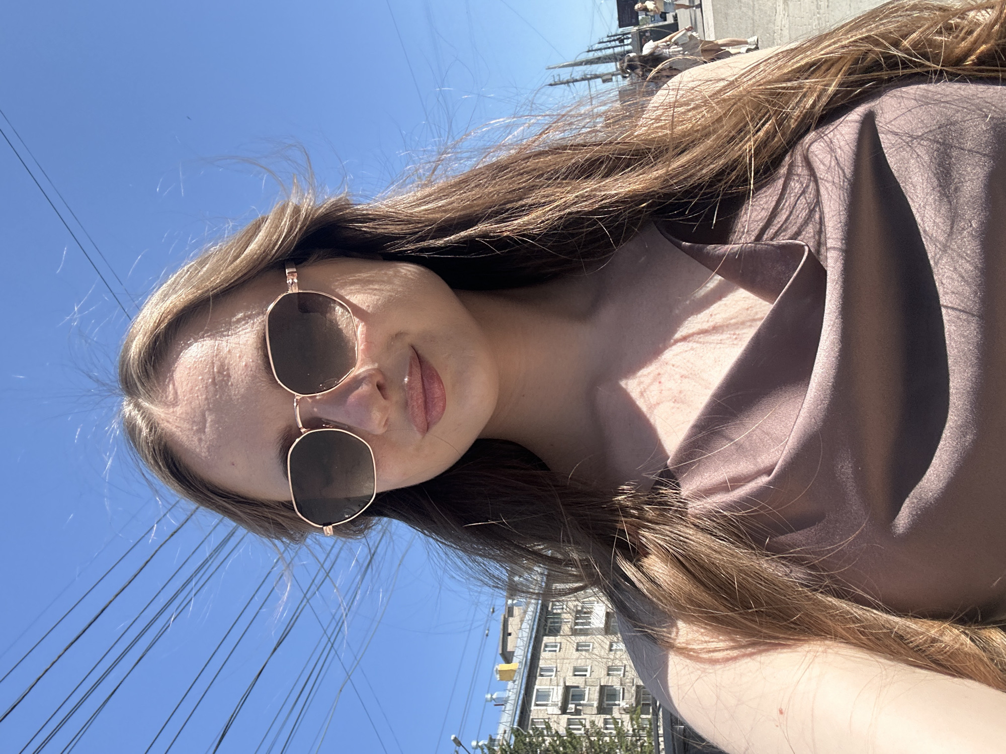 A young woman with long, brown hair and sunglasses stands on an outdoor city street on a sunny day, looking at the camera. She is wearing a draped, taupe-colored top. In the background, there are buildings, power lines, and other people walking. The scene appears to be in an urban environment.