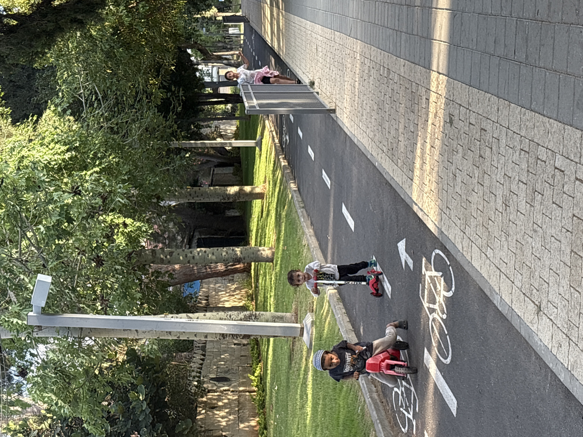 The image shows a paved path with white markings indicating a bicycle lane, located in the city of Holon, Israel. In the foreground, two young children are on the path. One child is riding a red toy motorcycle and is wearing a dark t-shirt with the word "FORTNITE" on it and a kufi hat. The other child is standing on a red scooter. Behind them, a slightly older child in a light-colored t-shirt and dark shorts is walking along the path, near a metal fence. The path is bordered by a strip of grass and then trees. The sun is casting shadows, suggesting it is either morning or late afternoon. The overall scene depicts a recreational area within a city.