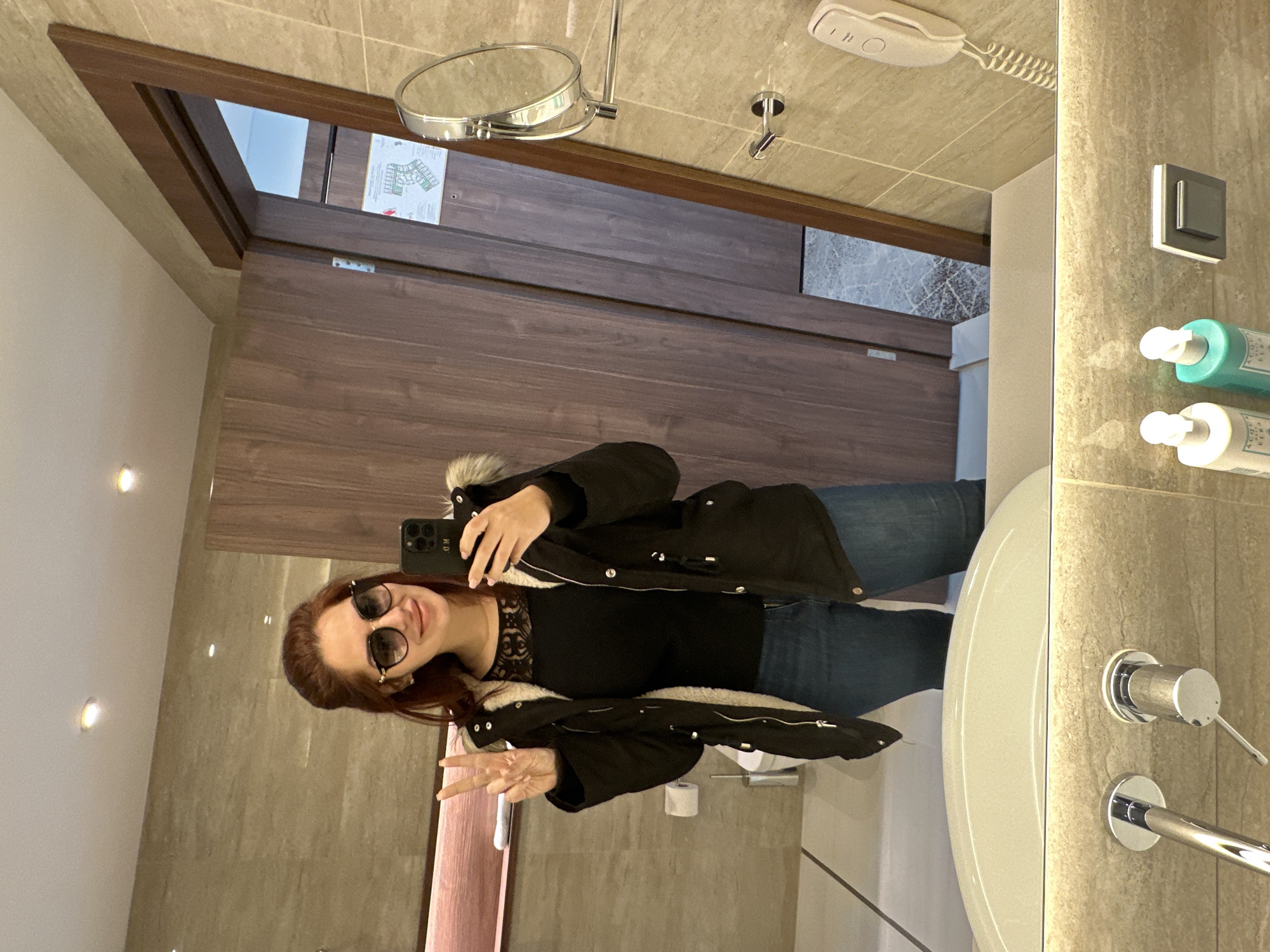A young woman is captured in a mirror selfie inside a modern bathroom, likely within a hotel or apartment setting, potentially in Kazanlak, Bulgaria. She has reddish-brown hair and is wearing sunglasses, a black lace-detailed top, blue jeans, and a black winter coat with a faux fur-lined hood. She holds a dark-colored smartphone, which has "MD" visible on its case, in her right hand to take the photo. Her left hand is raised, making a peace sign, and she displays a happy, content smile.
The bathroom features light beige tiled walls and floor, a white vanity with a sleek sink, a modern chrome faucet, and various fixtures including a wall-mounted magnifying mirror, a white hairdryer with a coiled cord, and chrome towel rails. Two pump bottles, likely containing toiletries, are visible on a shelf to the right. Through the open wooden door reflected in the mirror, part of another room is visible, including a wall with a diagram (possibly a fire escape plan or hotel layout) and hints of a light-colored ceiling with recessed spotlights. The overall setting is clean and contemporary, with ample indoor lighting. The woman's winter attire suggests the possibility of cold weather outside.