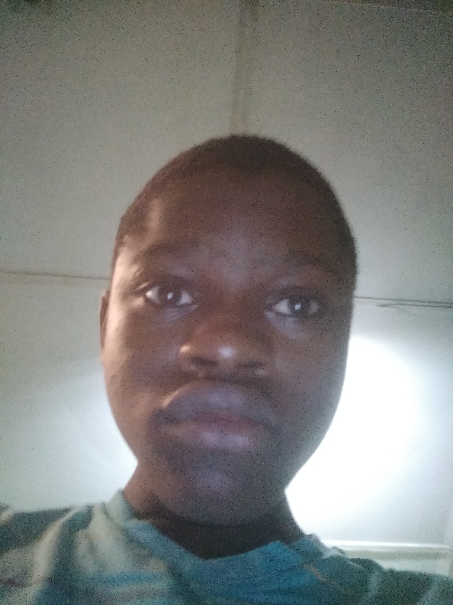 A medium close-up shot shows a person with dark skin, gazing directly at the camera. The person has close-cropped hair, dark eyes, a wide nose, and full lips. They are wearing a teal shirt. The background is a blurred, off-white surface. There is no indication of location.