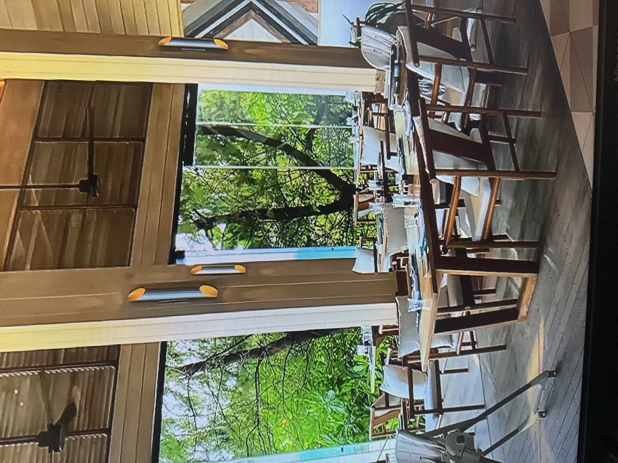 The image depicts the interior of a modern, upscale restaurant with large windows overlooking lush greenery. The restaurant features wooden tables and chairs with white cushions, creating a clean and elegant aesthetic. Pendant lights with warm, orange glows are mounted on the wooden beams, adding to the ambiance. Ceiling fans are visible, suggesting a climate-controlled environment. The overall scene conveys a sense of tranquility and sophisticated dining. There is no visible indication of the restaurant being located in Kyiv, Ukraine.