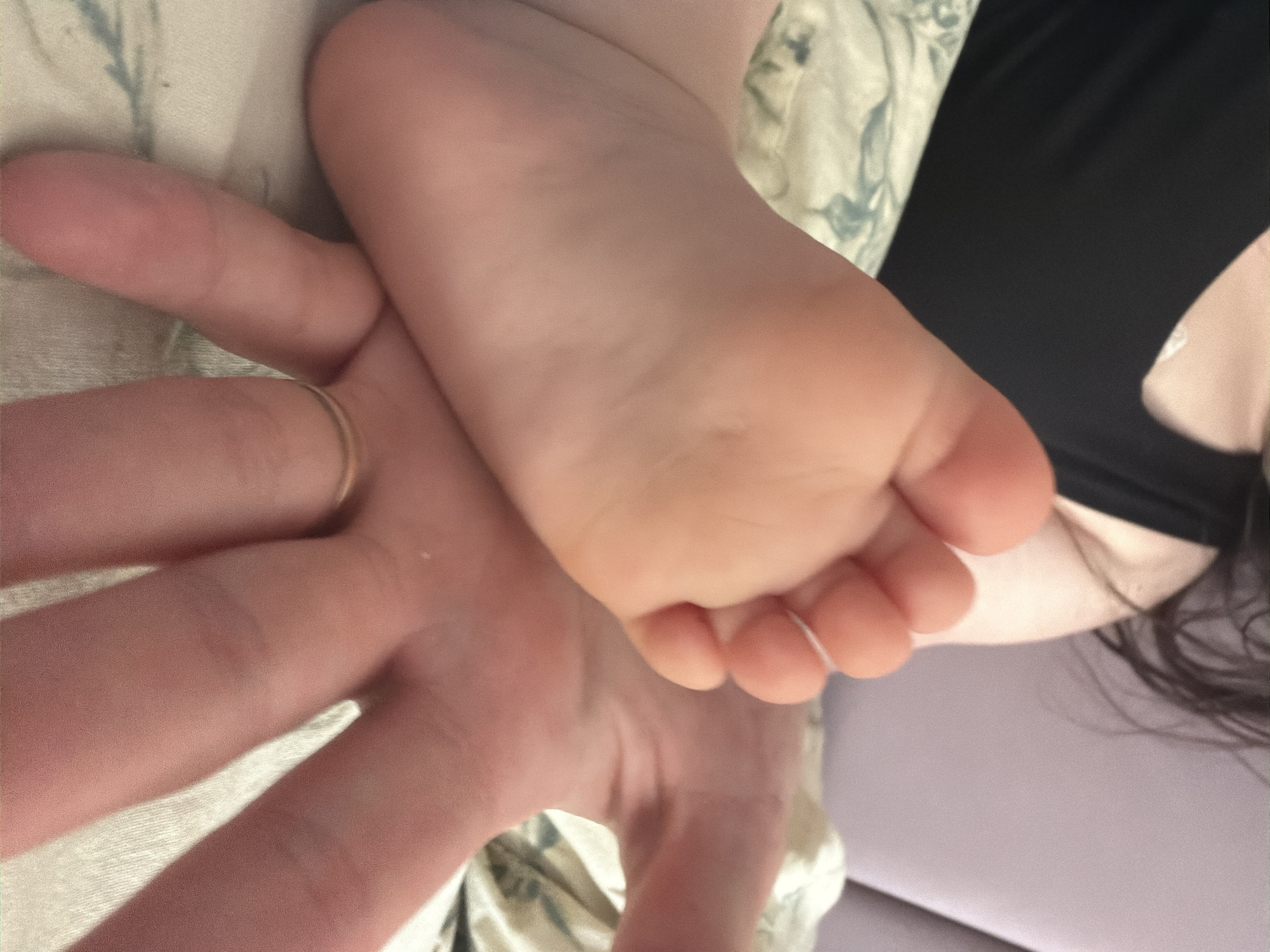 A close-up photograph shows a person's hand with a gold ring on one finger holding a baby's foot. The person is wearing a black tank top and lying on a bed with a patterned pillow. The background includes a lavender-colored surface. There is no discernible information to suggest that the location is Staraya, Russia.