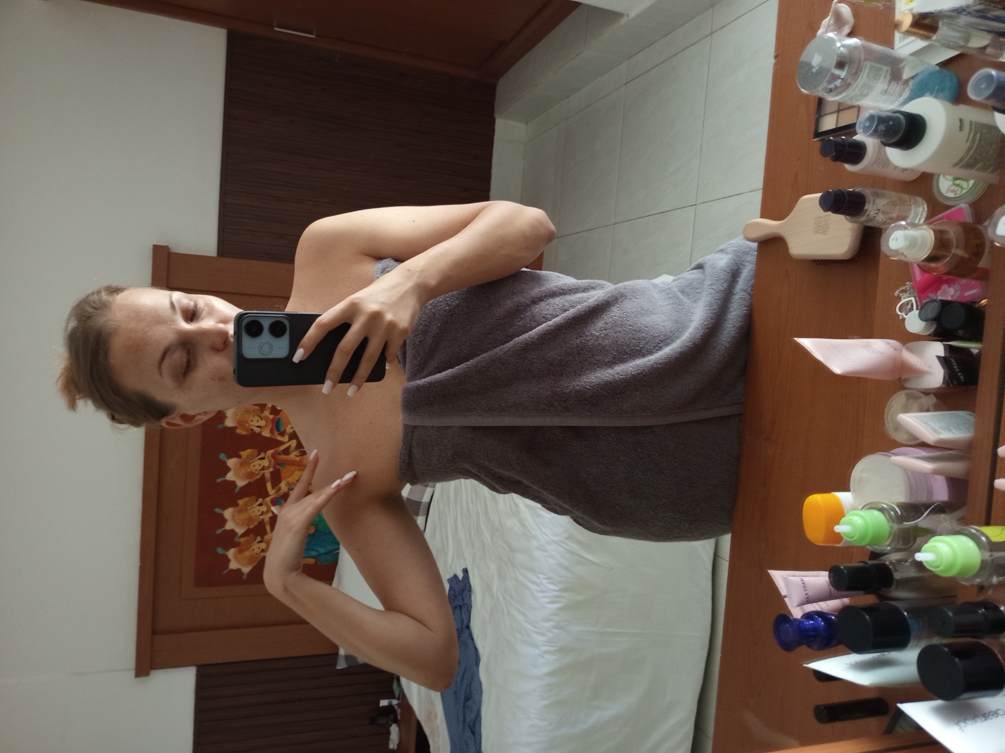 A woman is standing in front of a mirror, taking a selfie. She is wearing a gray towel wrapped around her body and has her hair pulled up in a bun. The room appears to be a bedroom, with a bed in the background and a vanity table in the foreground. The vanity table is cluttered with various cosmetic products. On the wall behind the bed, there is a decorative artwork depicting several figures. The floor is tiled. The overall impression is that of a private moment in a hotel room in Denpasar, Indonesia, possibly after a shower.