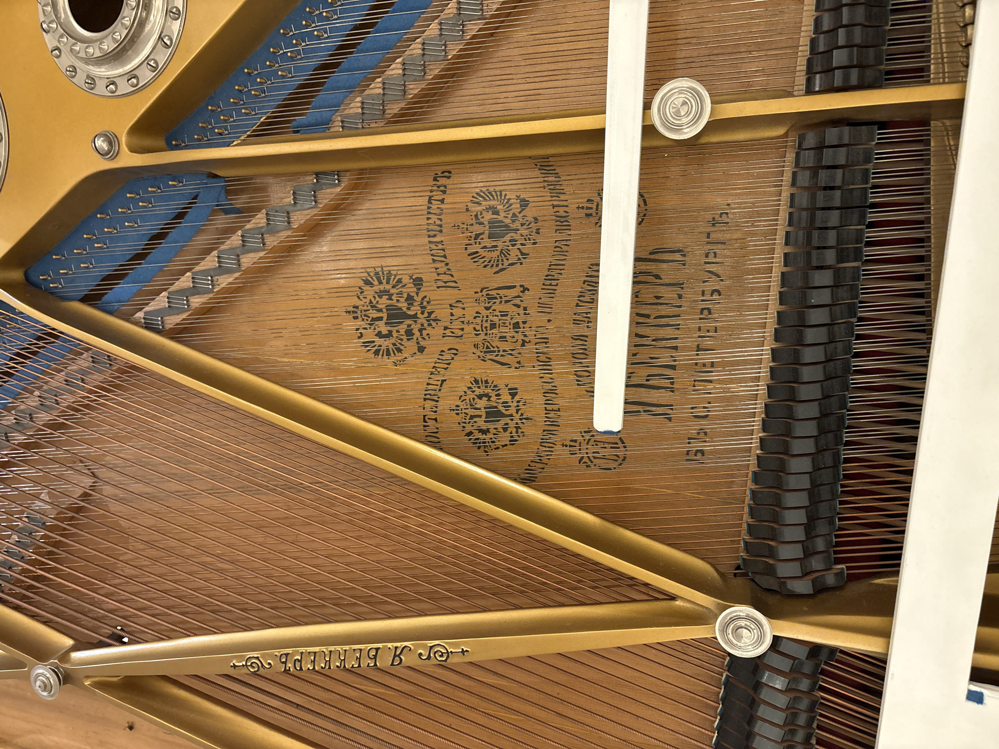 This media file captures a detailed, top-down view of the internal components of a grand piano. The golden-colored metal frame, which provides structural integrity, is prominently featured, contrasting with the warm, light-brown wooden soundboard beneath. Numerous piano strings, some thinner and silver-colored for higher notes and others thicker and copper-wound for bass notes, stretch across the soundboard in parallel lines.
On the left, multiple tuning pins are visible, embedded in blue felt strips, securing the strings. On the right side, a series of black felt dampers rest upon the strings, arranged in a stepped pattern. Several silver, circular agraffes are also visible, guiding and spacing the strings.
The wooden soundboard displays intricate decorative patterns and Cyrillic text stamped into its surface. Key visible text includes:
- "ПОСТАВЩИКЪ ДВОРА ЕГО ВЕЛИЧЕСТВА" (Supplier to His Majesty's Court), indicating a prestigious status.
- "ВЪДАЛЬНЪИШЕЕ" (V'dal'nyshee), likely part of a regal honorific phrase.
- "ВЪ САНКТЪ-ПЕТЕРБУРГЪ" (In Saint Petersburg), suggesting the place of origin or significant association.
- The manufacturer's name, "Я. БЕККЕРЪ" (Ya. Bekker), appears inverted on the lower golden frame and partially visible on the soundboard.
The image provides a close-up of the instrument's complex internal mechanics and historical craftsmanship. There is no specific activity taking place, as it is a static shot of the piano's interior. The setting is indoors, with no indications of time of day or weather. The location context provided is Khimki, Russia.