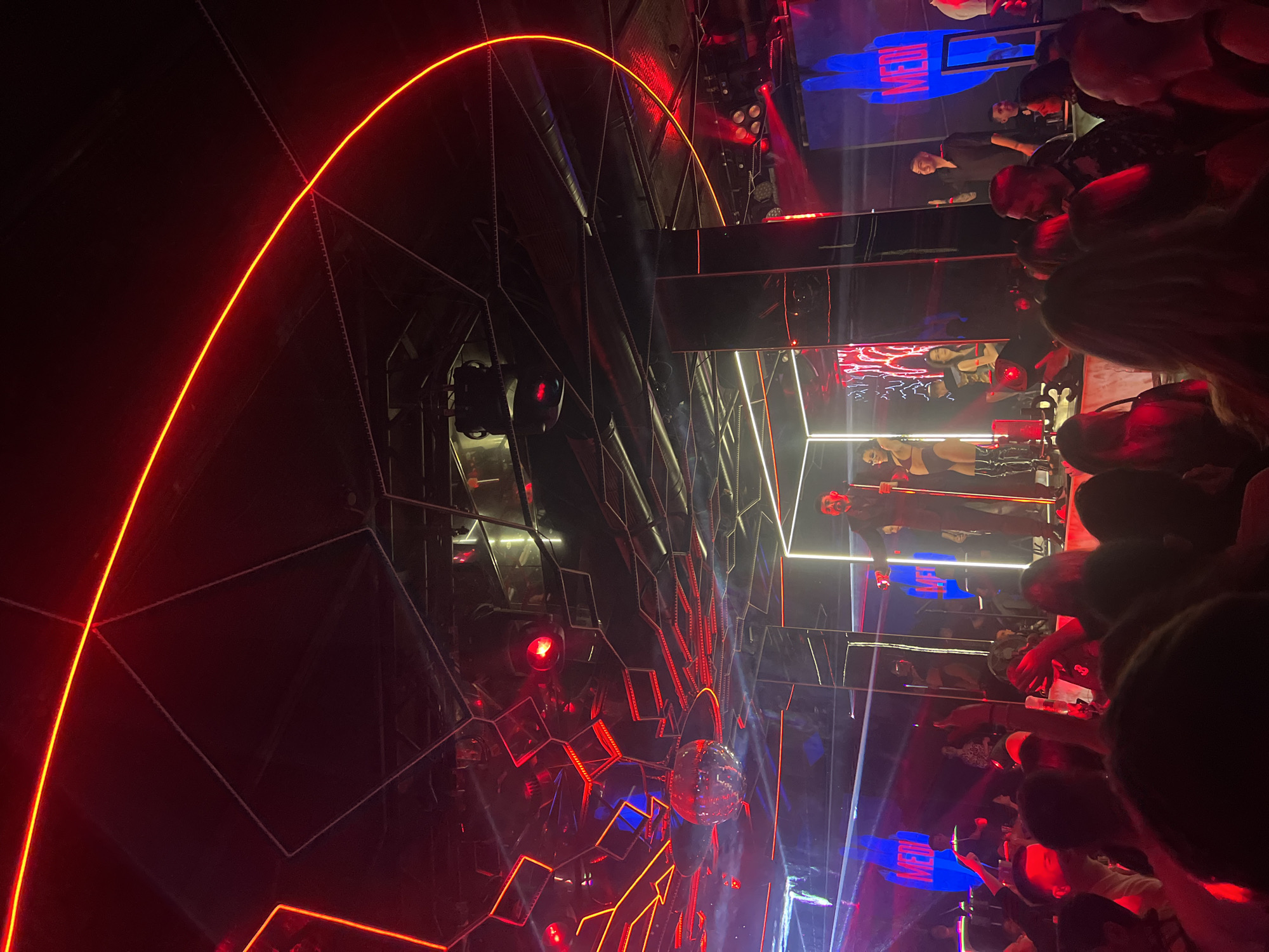 This image depicts a live performance within the interior of a crowded nightclub, identified as Club MEDI in Sofia, Bulgaria. The scene is illuminated by red and blue artificial lighting, including neon strips and spotlights.
A male performer, likely a singer or DJ, stands centrally on a stage, holding a microphone in his right hand. He is dressed in dark attire, possibly a suit. To his left, a female dancer in a dark, two-piece outfit and thigh-high boots poses with her left arm raised. Another female dancer is partially visible to the right. Behind the performers, large vertical LED screens display graphics, prominently featuring the word "MEDI" in white against blue and red backgrounds.
The club's ceiling is dark and features a geometric pattern of red neon strip lights, including a large curved line and several angular shapes. A reflective disco ball hangs from the ceiling. Multiple reflective pillars and surfaces are integrated into the stage design and surrounding environment, reflecting the vibrant lighting.
A dense crowd of individuals occupies the lower half of the image, facing the stage. Their heads and shoulders are visible, with some arms raised, suggesting engagement with the performance. Several individuals are holding drinks. A male individual on the right side of the stage, separate from the main performer, is seen smiling and waving towards the audience.
