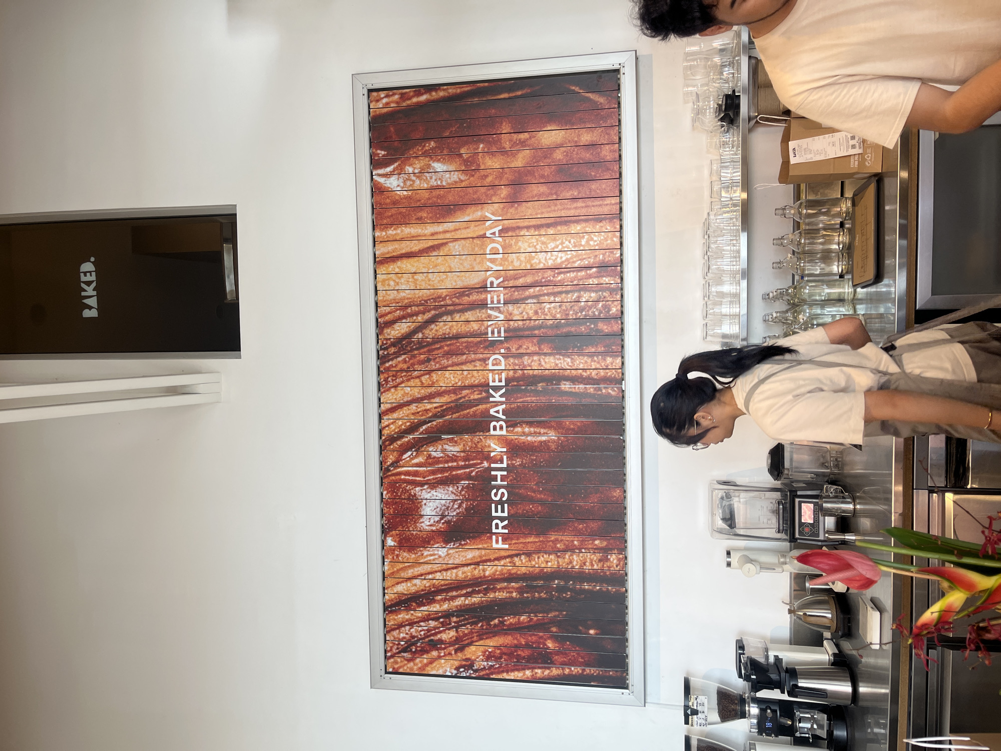 A brightly lit commercial interior shot in North Kuta, Indonesia, shows a service area of a bakery or café. Two individuals, possibly employees, are seen working behind a stainless steel counter. One person with dark hair and a white headband is leaning over the counter, while another individual in a white t-shirt is standing upright beside them. Behind the counter, shelves are stocked with glassware and containers. Equipment such as blenders and coffee grinders are visible on the counter. A floral arrangement in red and yellow is positioned at the front of the counter. On the left wall, a dark rectangular sign with the word "BAKED." in white is mounted above a horizontal shelf. Next to it, there is a framed graphic panel. The panel displays a close-up image resembling baked goods or bread arranged in horizontal stripes, with the words "FRESHLY BAKED. EVERY DAY" vertically printed on the graphic.