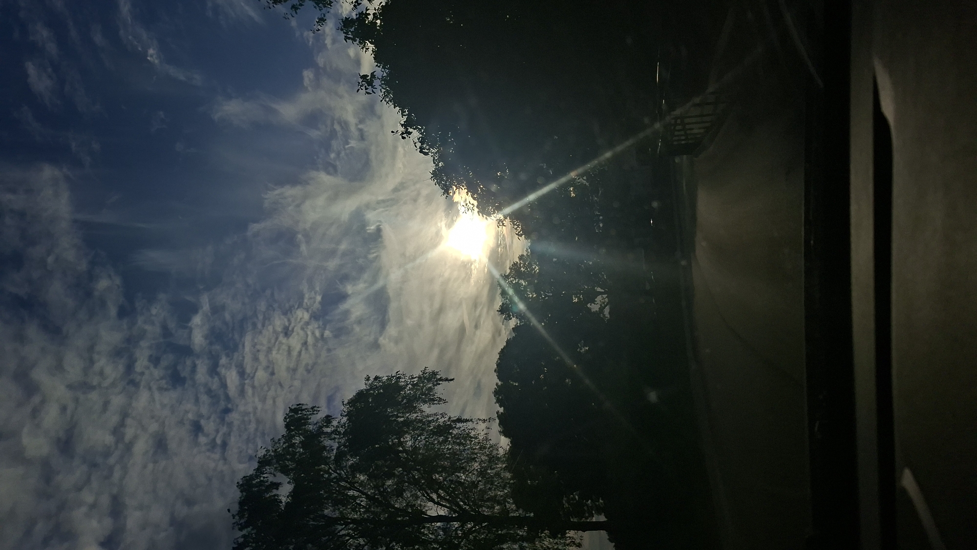 The image shows a view of the sky and trees from inside a vehicle, possibly on a road. The sun is visible, partially obscured by clouds and trees, casting light rays. The sky is a deep blue with scattered white clouds that have a wispy, feathery appearance. Silhouetted trees line the sides of what appears to be a road leading into the distance. The foreground shows the dashboard of the vehicle. There are no people or specific landmarks identifiable that would pinpoint the exact location within Smalyavichy, Belarus. The overall scene suggests a natural setting at daytime, possibly late afternoon or early evening due to the sun's position and the contrast between light and shadow.