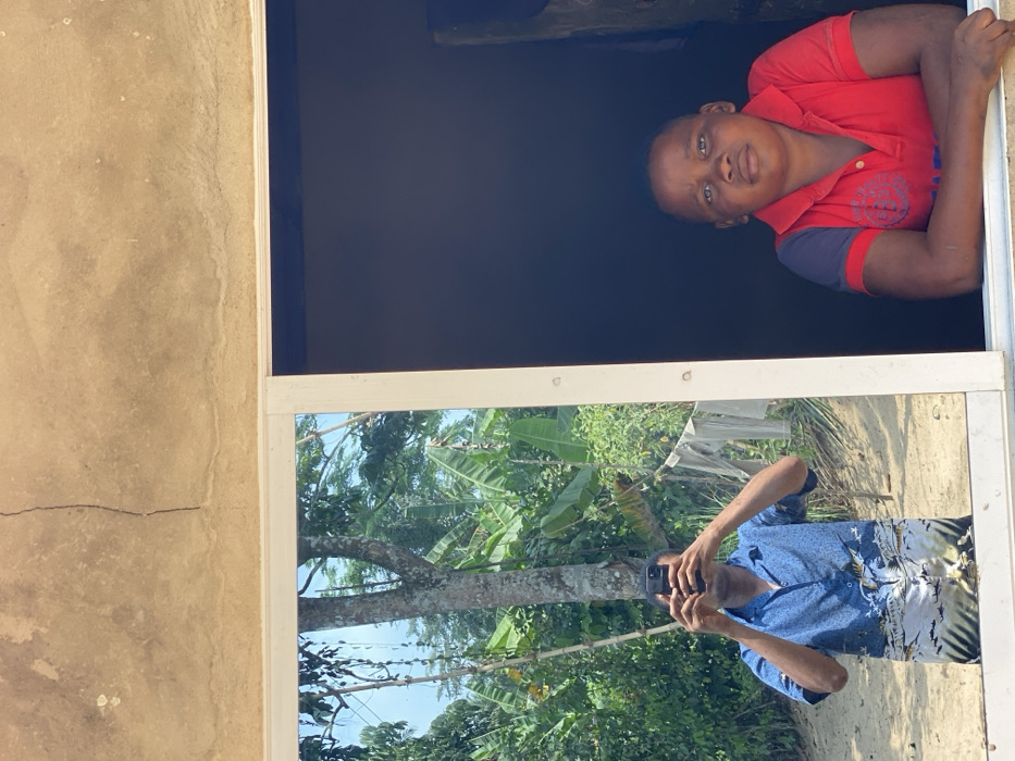 The image depicts a scene observed through and around a window in Ikot Ete Ekpo, Nigeria. The upper portion shows a light brown concrete wall with a prominent vertical crack and minor surface imperfections. Below the wall is a white-framed window, divided into two panes.
The left pane is reflective, mirroring an outdoor environment. Reflected in this pane is an individual, likely an adult male, holding a black smartphone vertically with both hands, appearing to be taking the photograph. He is wearing a blue short-sleeved collared shirt with a patterned lower section. The reflected outdoor scene includes a large tree with light-colored bark, dense green foliage consistent with banana plants, and patches of sandy ground under bright daylight. White or light-colored laundry is visible hanging on a line or bush in the reflected background.
The right pane of the window opens to a dark interior. From this interior, a young male looks directly towards the viewer, resting his forearms on the white windowsill. He is wearing a red short-sleeved polo shirt with dark blue trim on the sleeves. His expression is neutral.
