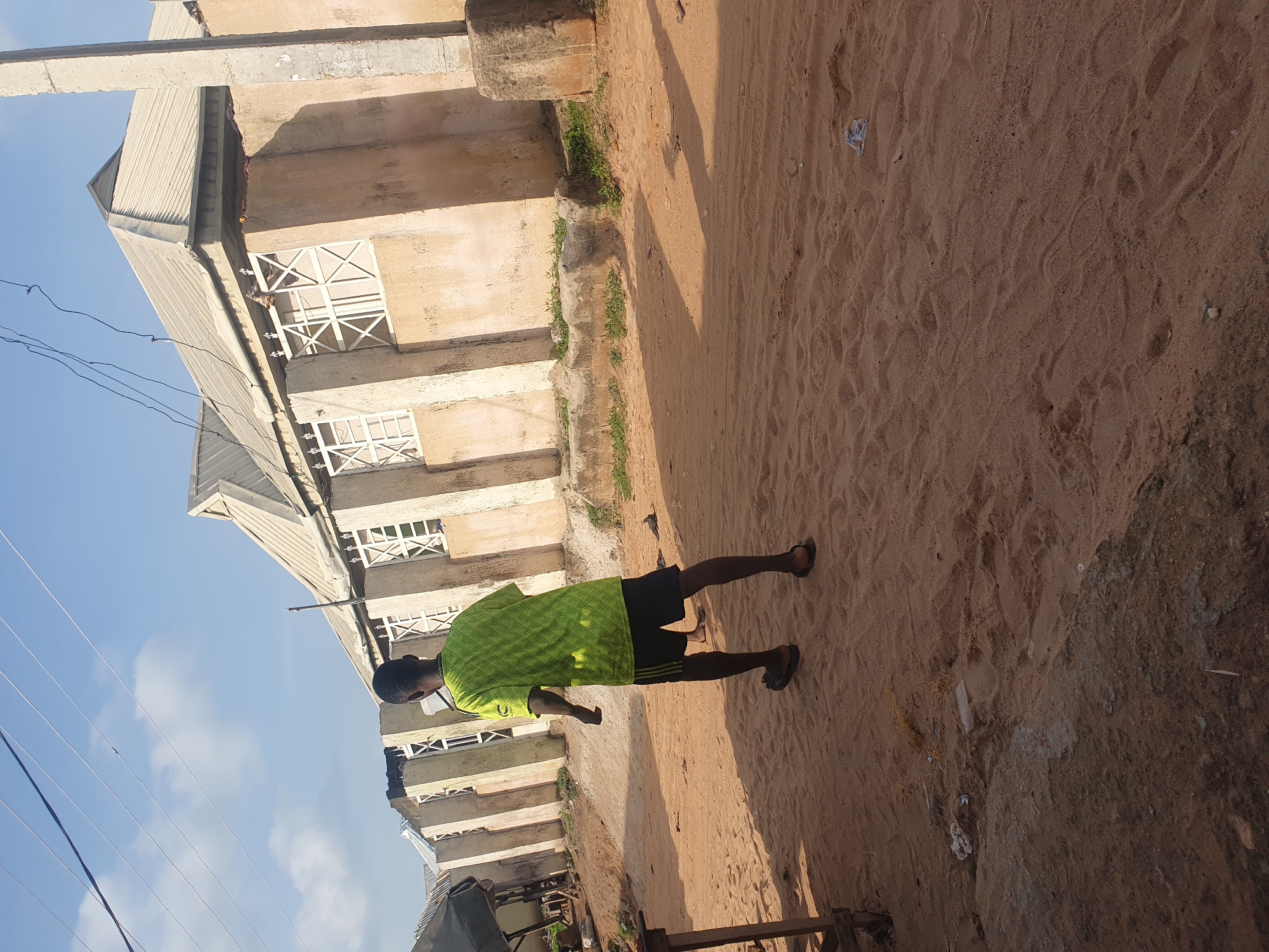 A person is standing on a dirt ground, facing away from the camera and towards a building. They are wearing a bright green shirt and black shorts, with sandals on their feet. The building appears to be under construction or unfinished, with exposed concrete and metal framing visible. The ground is covered in sand and some sparse vegetation. Overhead, power lines crisscross against a clear blue sky. The sun is casting shadows, indicating it is likely daytime. The overall scene suggests an outdoor urban or semi-urban environment, possibly in a developing area. There is no visible text in the image.