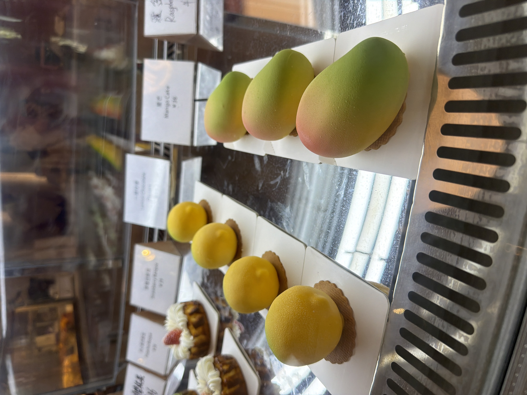 This image captures a display case showcasing an assortment of desserts, likely in a bakery or cafe in Yuzhong District, China. The scene is dominated by two rows of artfully crafted pastries.
In the upper row, three elongated, mango-shaped desserts are prominent. These pastries exhibit a smooth, matte finish with a striking color gradient, transitioning from green at one end to yellow in the middle, and finally to a subtle reddish-pink at the other end. Each sits on a small, light-colored tart or cookie base. A visible white label to their left clearly reads "Mango Cake" in English, with corresponding Chinese characters above it (likely "芒果蛋糕"), and a price of "¥38" below.
The lower row features four spherical, vibrant yellow pastries. These desserts have a textured, almost frosted appearance, resembling lemons. They are presented on individual scalloped, brown bases, possibly made of shortbread or a similar pastry. Further down this shelf, another type of dessert is partially visible, appearing to be a mini Bundt cake or fluted pastry adorned with a dollop of white cream and a red strawberry.
Other white labels are scattered throughout the display. One further up in the display case shows "Raspberry" in English with Chinese characters. Another partially visible label mentions "Strawberry Banana."
The display case itself is made of reflective glass, showing blurred reflections of the interior environment and a faint, distorted reflection of a person in the left-hand panel. The overall presentation is clean and organized, designed to showcase the desserts appealingly. The combination of English and Chinese text on the labels indicates the location as a Chinese-speaking region catering to both local and international customers.
