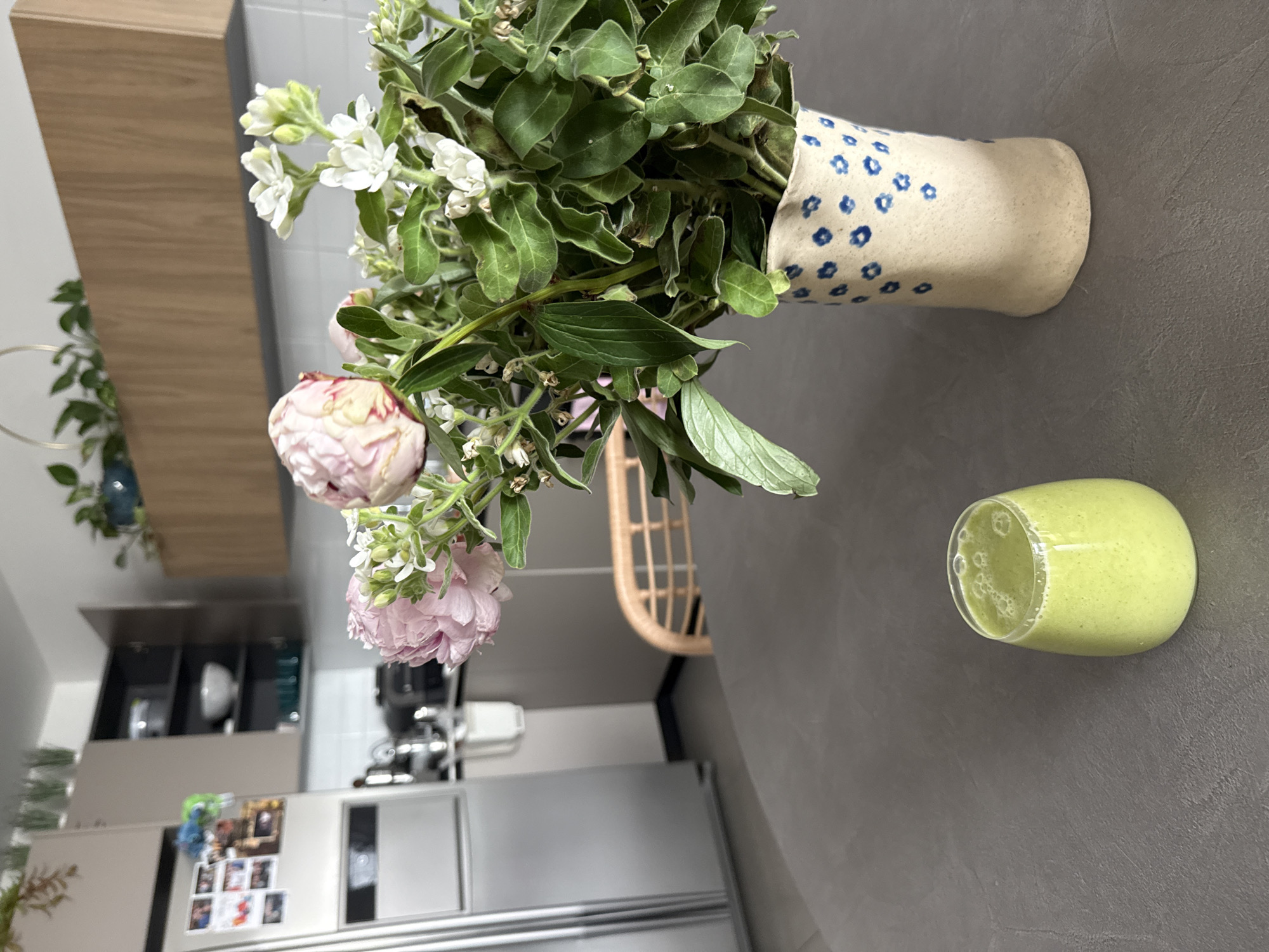A still life scene is positioned on a dark gray countertop. A vase with blue floral patterns holds a bouquet of flowers featuring pink peonies and smaller white blooms amidst green foliage. Adjacent to the vase, a glass of green smoothie or juice sits. In the background, a modern kitchen is visible with stainless steel appliances and light gray cabinetry. The presence of magnets and photos on the refrigerator suggests a domestic setting. The location cannot be determined based on the image.