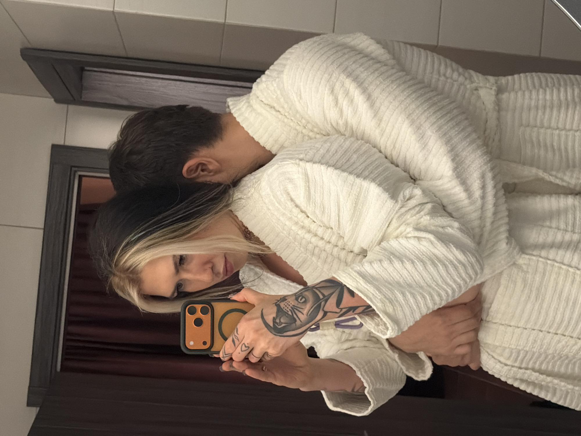 A woman with dark and blonde hair and a tattoo on her arm is holding a yellow phone and taking a selfie in front of a mirror, embracing a person in a white robe. The location appears to be a bathroom setting with tiled walls and a dark-framed mirror. There is no information to determine if this image was taken in Palilula Urban Municipality, Serbia.