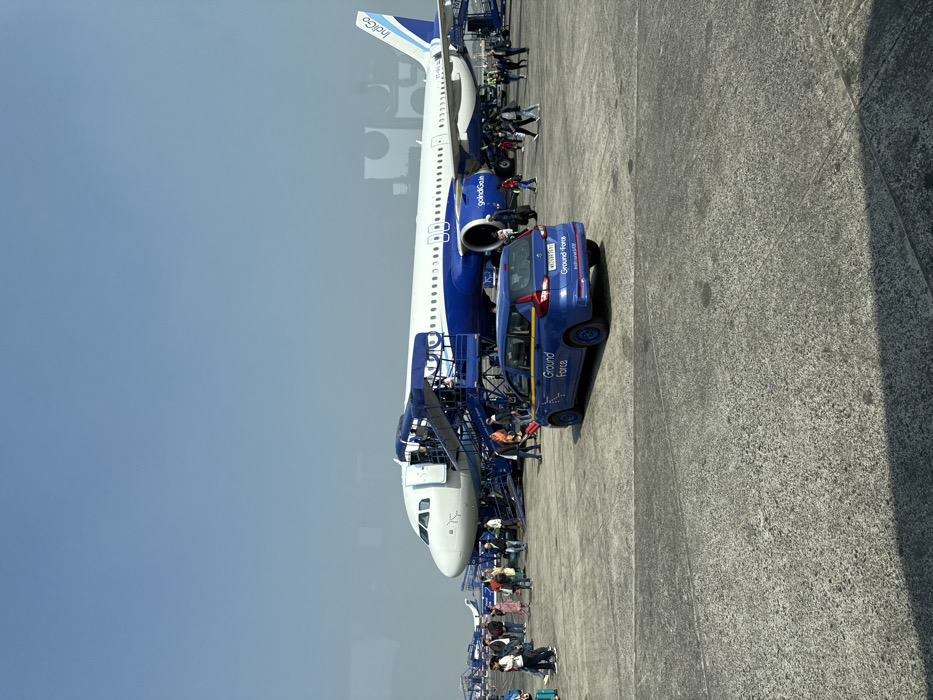An Indigo airplane is parked on the tarmac, with a blue "Ground Force" vehicle nearby. Passengers are boarding or deplaning via a mobile staircase attached to the aircraft's front door. The sky is clear, and a concrete surface surrounds the plane, showing signs of wear. People are queuing, some with luggage, suggesting a typical airport arrival or departure scene.