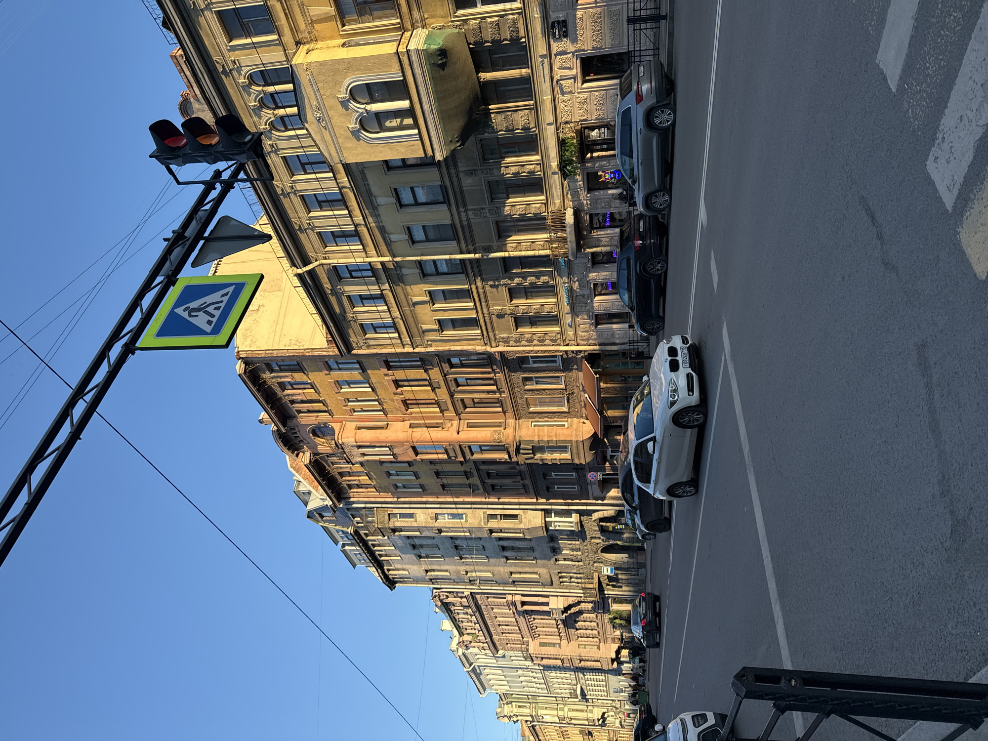 The image captures a vibrant, sunny street scene in Saint Petersburg, Russia, looking down a street lined with historic buildings. On the left, a row of ornate, multi-story buildings display varied architectural styles, with facades in shades of beige, grey, and reddish-brown, featuring numerous windows and some balconies.
The paved street on the right has several cars parked along the curb, including a prominent white BMW sedan with the license plate "T002EB 78" (where "78" indicates Saint Petersburg), a grey SUV, and other vehicles further down. A clear pedestrian crosswalk (zebra crossing) is visible on the asphalt. Overhead, a black metal pole supports a traffic light, which shows both red and amber lights illuminated, and a square blue sign indicating a pedestrian crossing with a white human figure.
The sky, visible in the upper left, is clear and bright blue. The warm, angled sunlight suggests it is either early morning or late afternoon, highlighting the architectural details and casting shadows. Small commercial signs are visible on the ground floors of some buildings, with text like "ФОРУМ" (FORUM) and "PЯC" faintly discernible. A black metal railing is partially visible in the bottom right foreground.