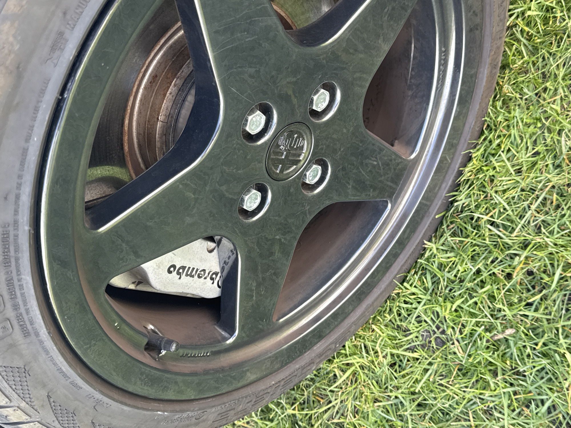 The image shows a car wheel and tire on grass. The wheel has a five-spoke design with a center cap displaying a logo. Five bolts secure the wheel. The tire is black and shows some wear. A Brembo brake caliper is visible behind the wheel. The scene appears to be outdoors, with green grass as the background. There is no information to place this specifically in Great Yarmouth, United Kingdom.