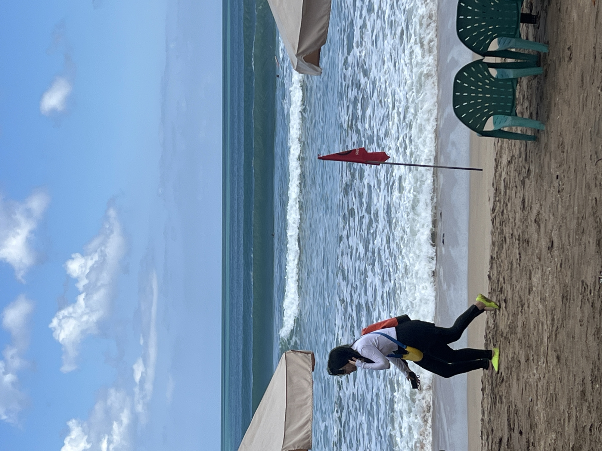 The image depicts a beach scene in Kuta, Indonesia, under a bright, partially cloudy sky. The ocean is a vibrant blue-green with moderate waves breaking along the shore, indicating surf conditions. In the foreground, a sandy beach is visible, with two empty green plastic chairs positioned on the right. A red flag with white lettering, likely a warning flag, is planted in the sand.
Two individuals are walking on the beach. The person on the left is wearing a white long-sleeved shirt, black pants, and bright yellow shoes. They have a yellow purse or bag hanging from their shoulder and are carrying a phone in their left hand. The second person, slightly behind and to the right, is also wearing black pants and bright yellow shoes. They are wearing a white long-sleeved shirt, and an orange bag is visible over their shoulder. Both individuals appear to be walking away from the viewer, towards the right of the frame.
Two beach umbrellas, partially visible on the left and right edges of the image, provide shade on the sand. A few distant surfers or swimmers can be seen in the ocean. The overall scene captures a typical day at a popular Indonesian beach.