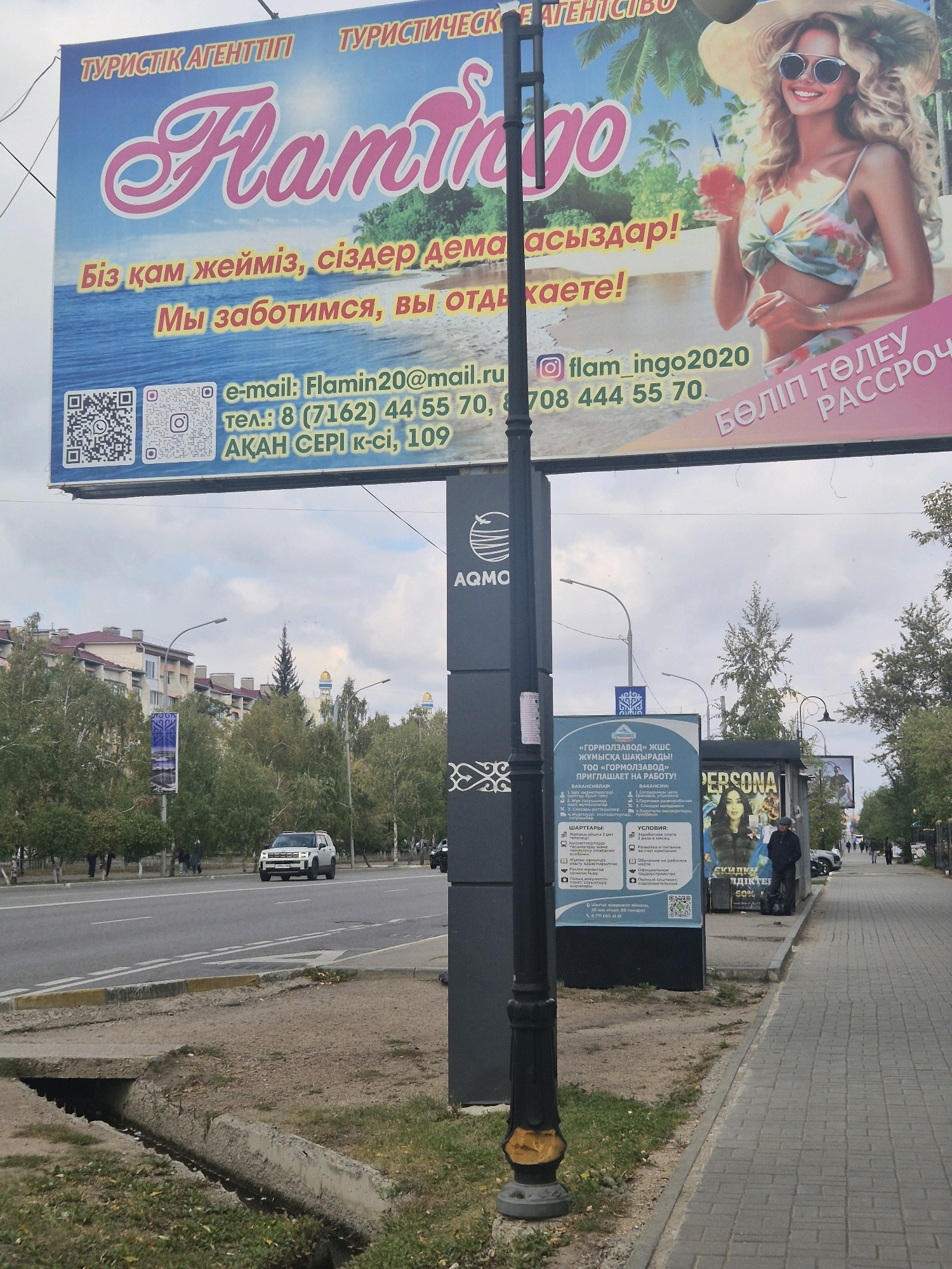 The image depicts a street scene in Kokshetau, Kazakhstan. A prominent billboard for a travel agency named "Flamingo" is visible, featuring a smiling woman in a hat holding a drink, set against a beach background. The billboard displays contact information, including an email address and phone number. Below the billboard, a tall, black lamppost stands near a sidewalk. To the right of the lamppost, a blue advertisement board lists job vacancies for "Gormolzavod," with details about qualifications and conditions. Further down the sidewalk, a kiosk displays a poster with the word "Persona" and a woman. The street itself is paved, with a white SUV driving on it. Buildings with red-tiled roofs and trees line the opposite side of the street. The sky is cloudy.