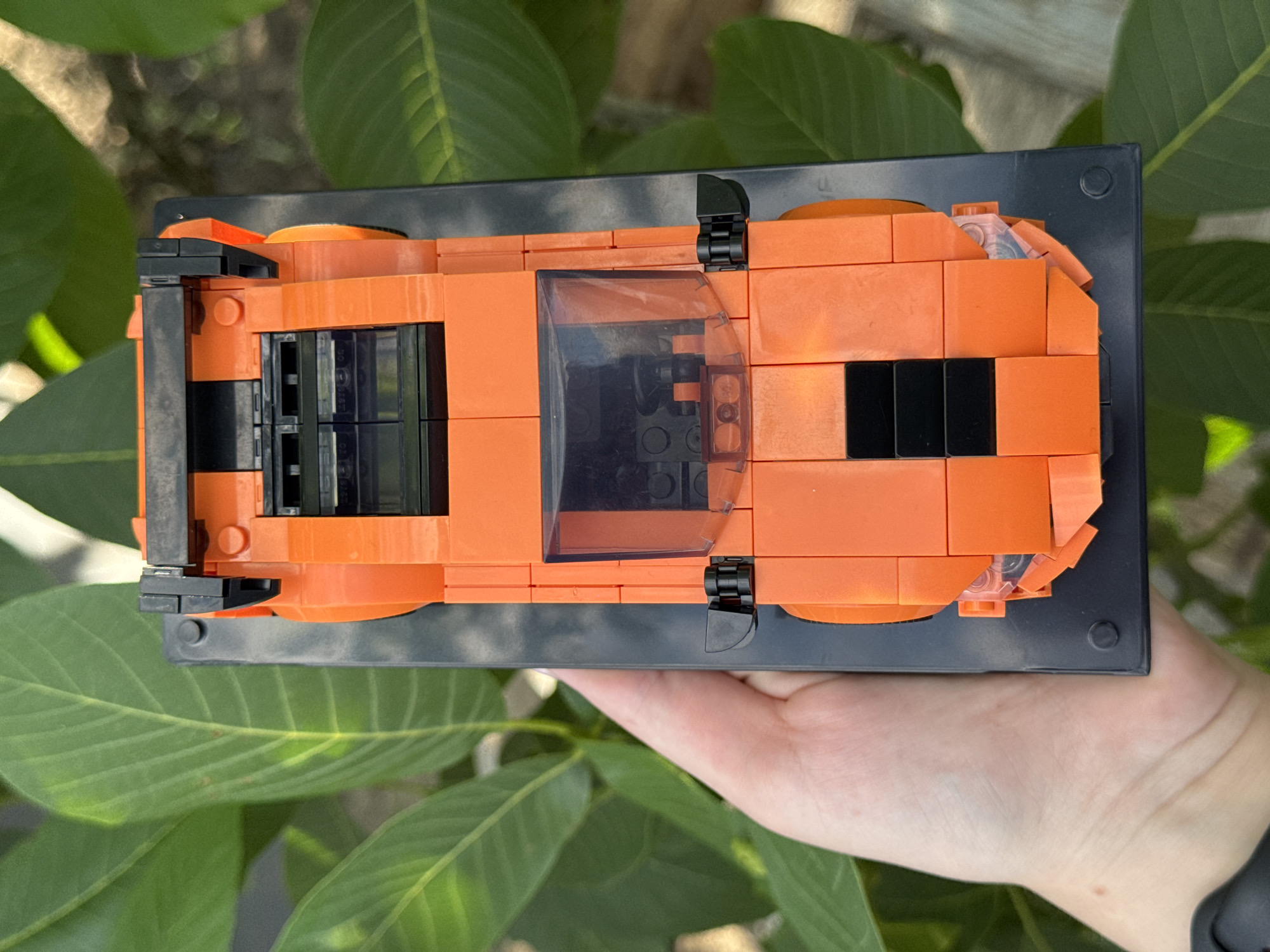 An overhead shot captures a bright orange Lego car with black accents, placed on a black Lego base. The car is positioned against a backdrop of lush green leaves, suggesting an outdoor setting. The scene is shot from a high angle, with a human hand gently holding the base of the Lego car. The car's details include a spoiler at the rear, a tinted windshield, and what appear to be headlights at the front. The overall impression is one of a playful, handcrafted object set against a natural environment.