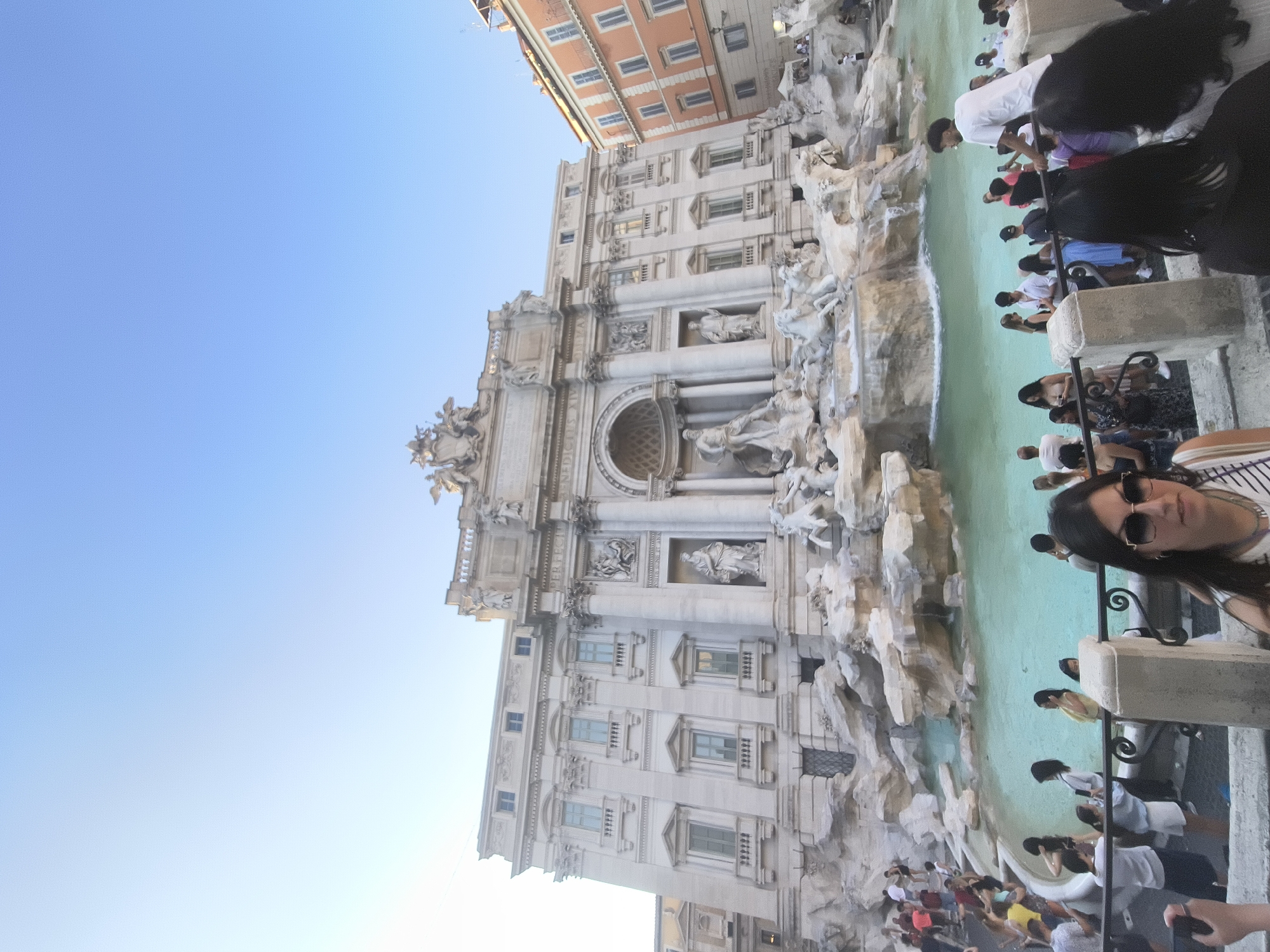 The image captures the Trevi Fountain in Rome, Italy, a popular tourist destination. The fountain is a large Baroque fountain, featuring a central statue of Oceanus. The water in the fountain is a vibrant turquoise color. A crowd of people are gathered around the fountain, many taking pictures and observing the sculptures. Some individuals are leaning on the railings, while others are standing closer to the water. The architecture surrounding the fountain is ornate and classical, with the Trevi Fountain itself being the main focal point. The sky above is clear and blue. The overall scene depicts a bustling tourist environment at a famous landmark.
