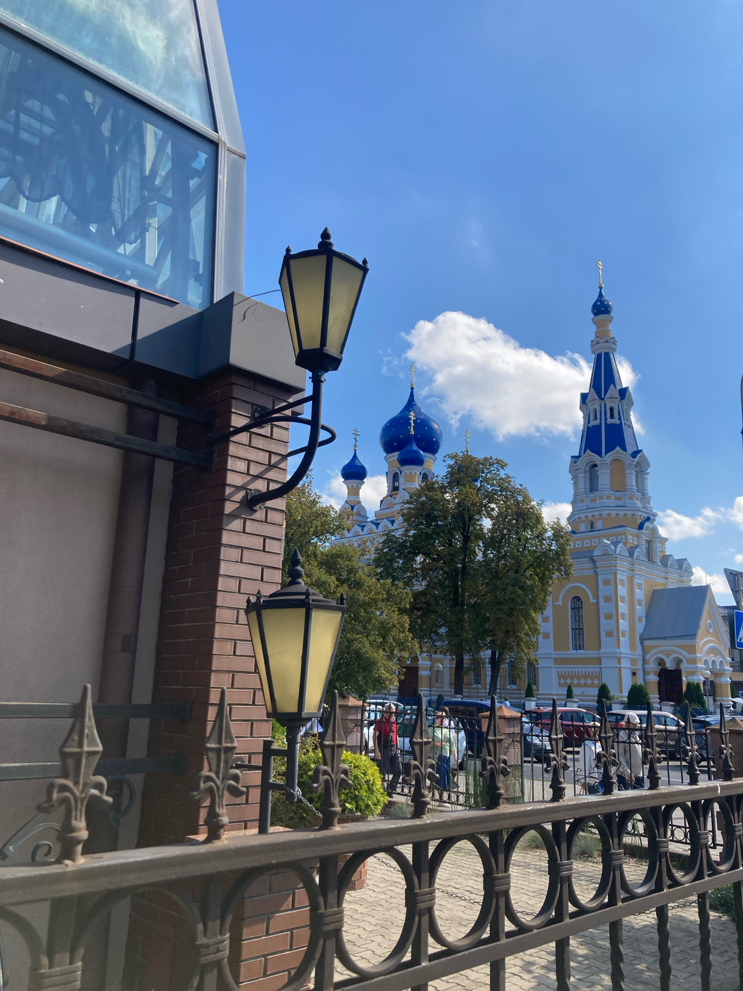 The photograph captures a view of the Holy Resurrection Cathedral in the city of Brest, Belarus, on a clear, sunny day with scattered clouds. In the foreground, a dark metal fence with decorative elements partially obscures the view. To the left, a modern building with a glass facade is visible. A brick pillar with two ornate lampposts is attached to the side of this building. In the middle ground, trees with green foliage are present. Beyond the trees, the yellow and white facade of the cathedral with blue domes and spires is prominently displayed, topped with golden crosses. Several cars are parked on the street in front of the cathedral, and a few individuals can be seen walking near the entrance. The ground is paved with light-colored cobblestones.