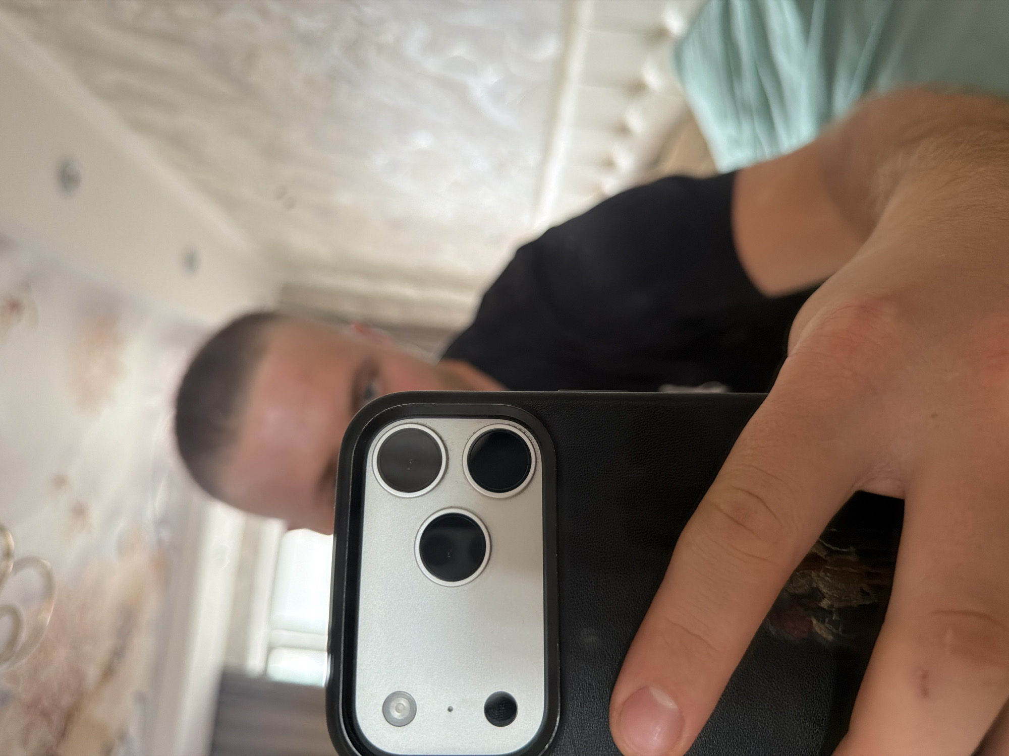 A close-up, low-angle, point-of-view shot captures a person holding an iPhone in their left hand, the phone's camera array in the foreground. The person is wearing a black t-shirt. The background is blurred, showing a white tufted headboard and a teal duvet, suggesting a bedroom setting. The ceiling above has light-colored wallpaper with a pattern and a light fixture. There are no visible landmarks or context to indicate a location in Strășeni, Moldova.