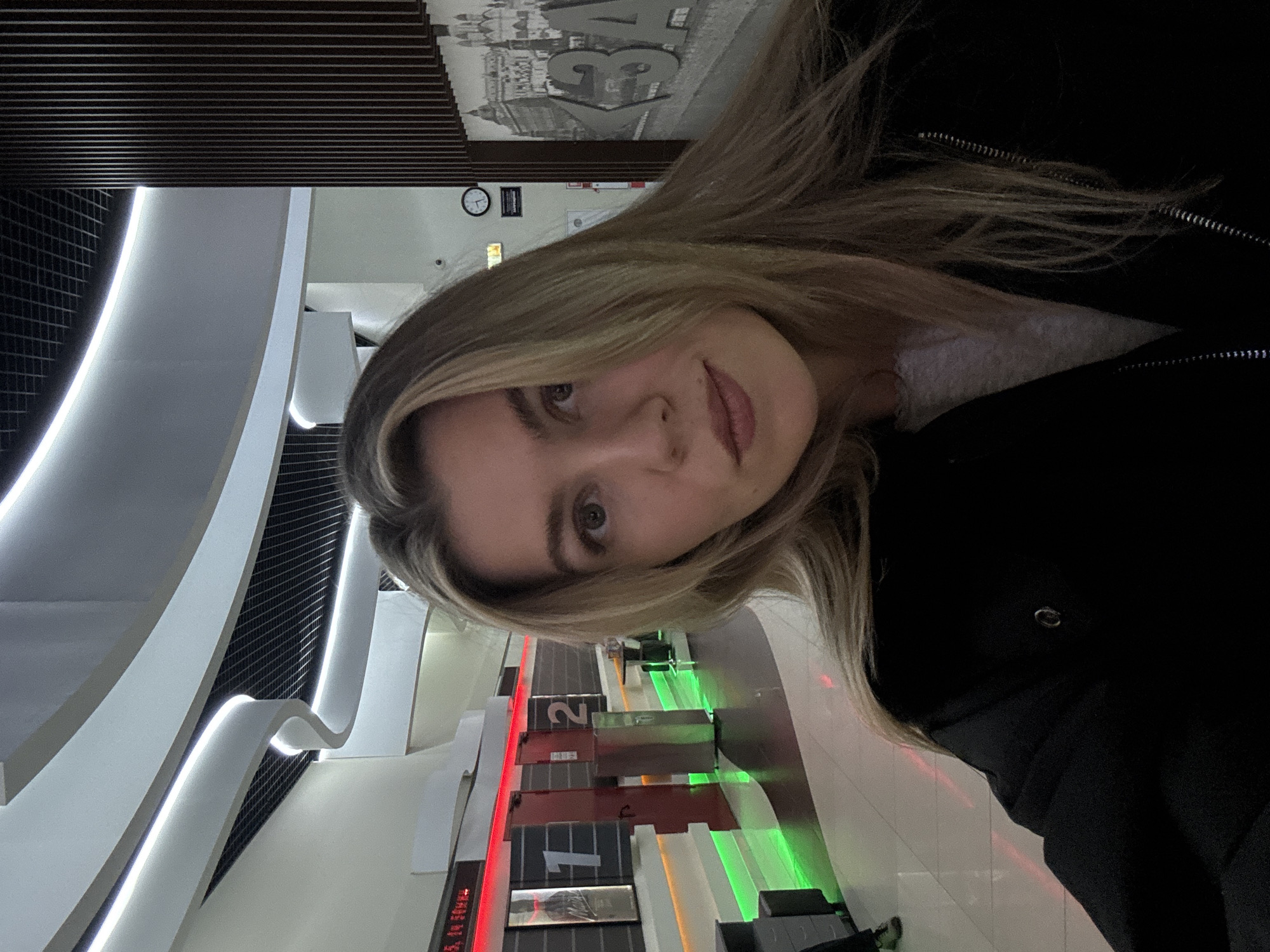A woman stands in a building with illuminated bowling lanes. The location is Unknown, Unknown. A woman with blond hair and a dark jacket is looking at the camera. Behind her are the bowling lanes lit up in green and red. White, wavy lighting is visible on the left.