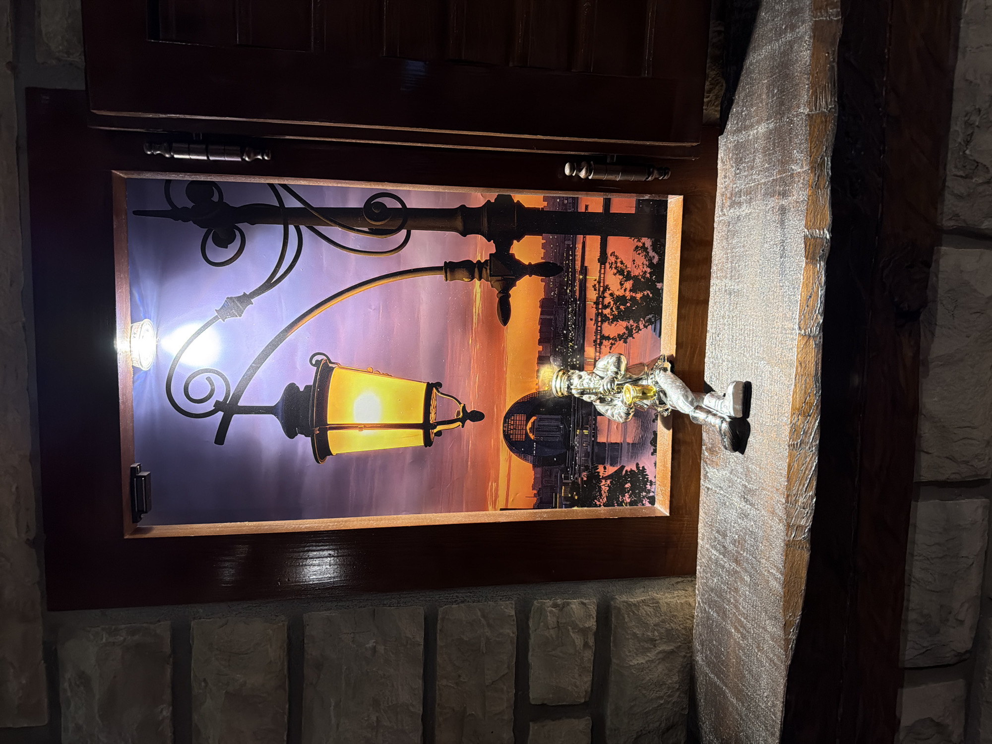 A small, silver figurine of a man playing a saxophone sits on a rough-hewn wooden ledge. The figurine is positioned in the foreground, facing left, with its legs dangling over the edge of the ledge. Behind the figurine, a framed picture depicts a city skyline at sunset, with a large, illuminated streetlamp in the foreground. The picture is housed within a wooden frame, which is slightly ajar, revealing a dark interior on the right. The ledge is set against a stone wall, with another section of stone visible below the ledge. The overall scene suggests an interior decorative display. The image does not provide any information that would indicate a location in Kumbor, Montenegro.