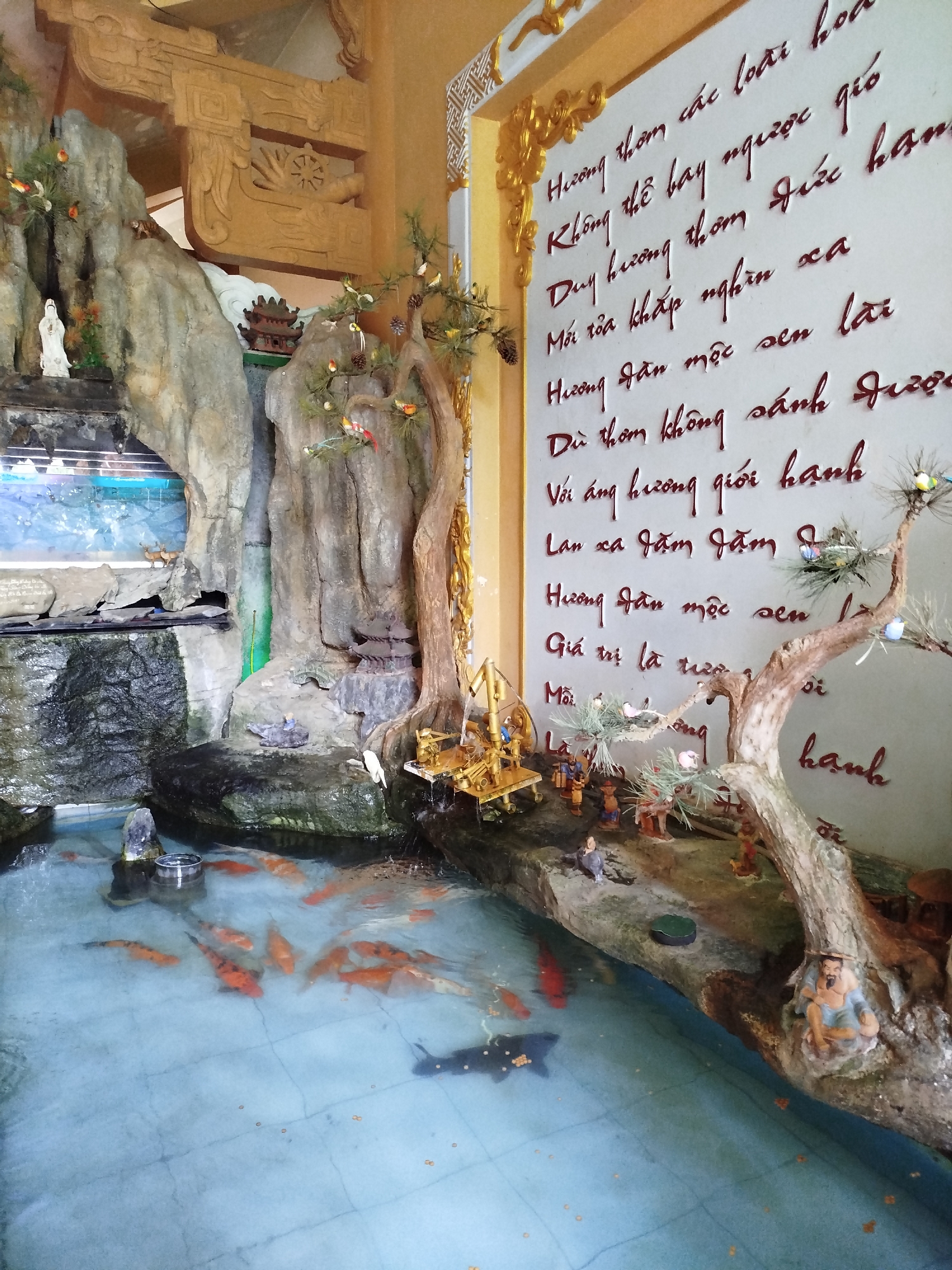 The image captures an indoor koi pond installation in Pleiku Ward, Vietnam. The pond is populated with numerous orange koi fish swimming in the water, interspersed with black fish. The pond’s backdrop consists of an artificial rock formation featuring a miniature waterfall and a small statue of a figure. On the right, the wall is adorned with inscribed text in Vietnamese, likely verses of poetry or scripture. Additional miniature figurines and trees are positioned along the edge of the pond, enhancing the artistic composition of the scene.