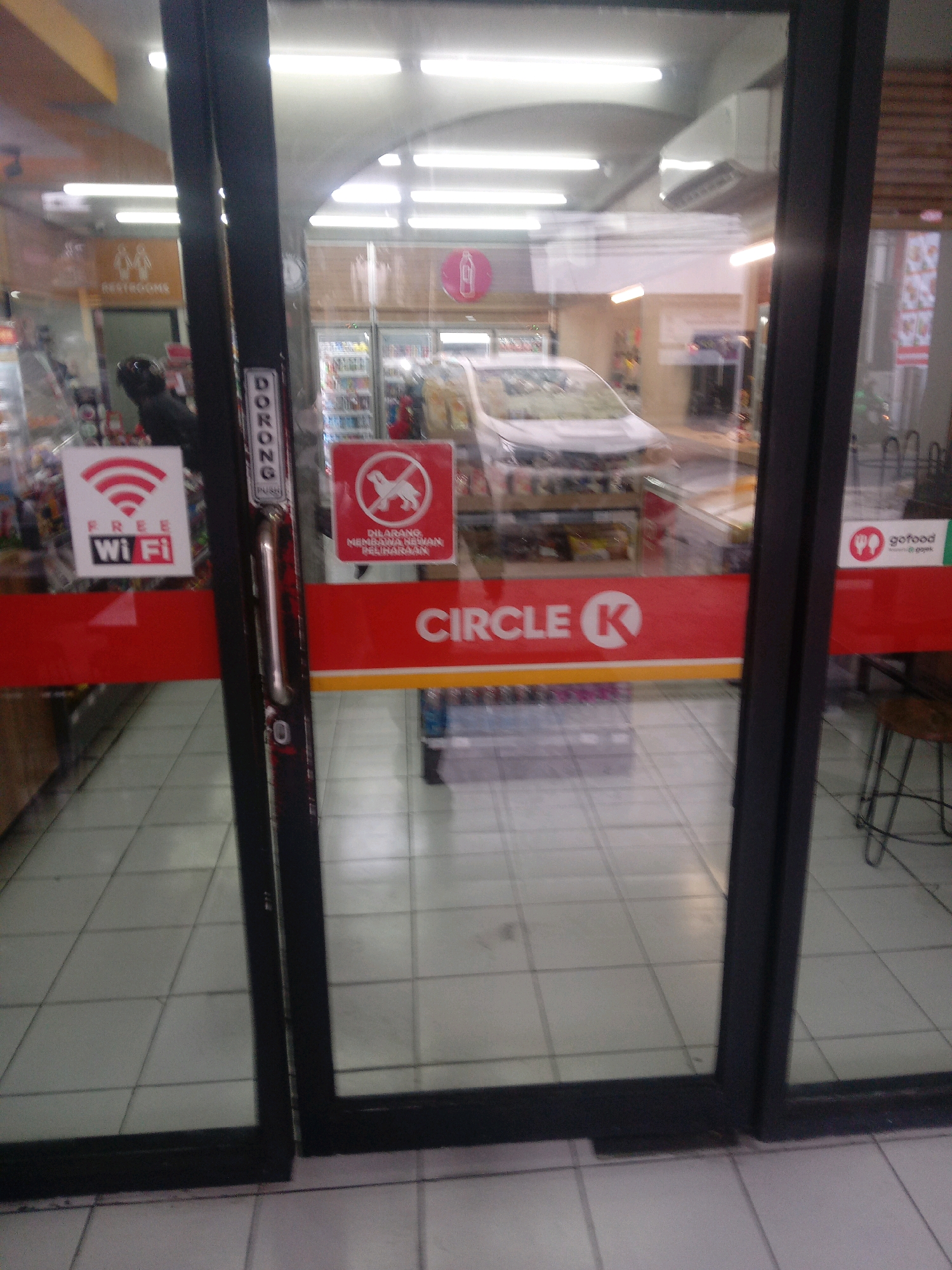 A medium shot captures the exterior of a Circle K convenience store in Depok, Indonesia, viewed through glass double doors. The doors are framed by black metal, and the floor is tiled with light gray squares. On the left door, a white sticker with red Wi-Fi signals and the text "FREE Wi Fi" is visible. Below this, a red band runs across the glass, displaying "CIRCLE K" and a red circle with a white "K" logo. On the right door, a red square sign with a white border depicts a dog silhouette with a red circle and slash through it, indicating "DILARANG MEMBAWA HEWAN PELIHARAAN" (No pets allowed). Further to the right, a red band also displays "CIRCLE K" and the logo, and a white sticker with red icons shows "gofood" and "@gojek." Inside the store, shelves stocked with goods and a white car are partially visible through the glass. The lighting suggests it is daytime.