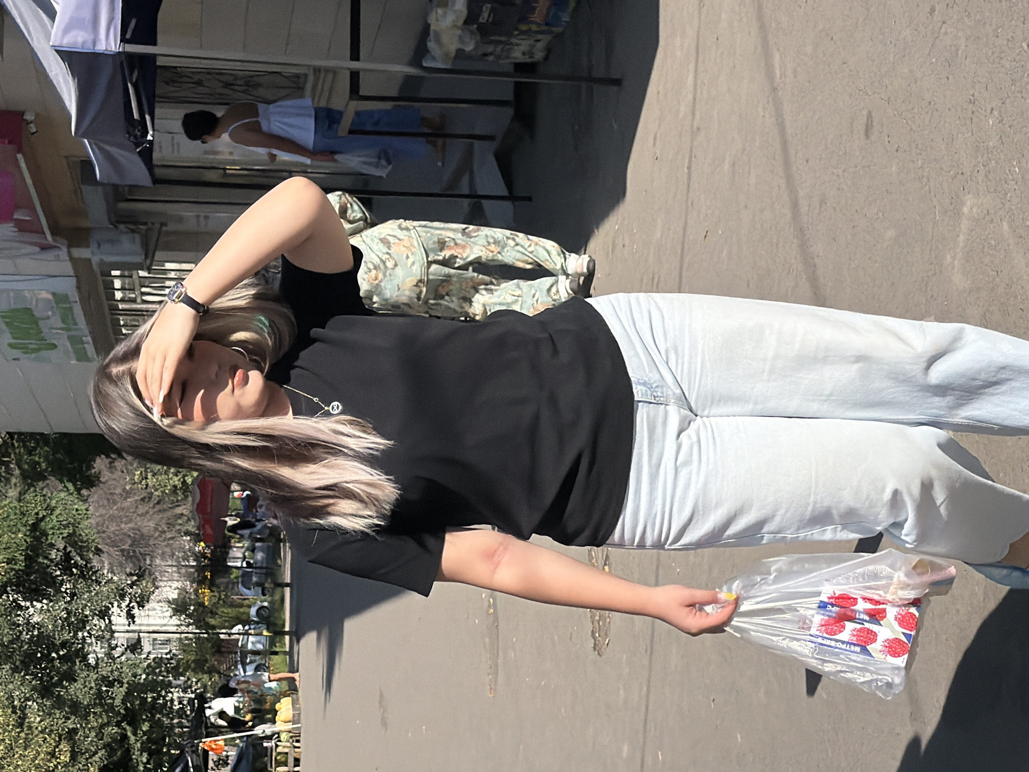A young woman with blonde hair, wearing a black t-shirt and light-wash jeans, walks down a sunlit street. She is holding a clear plastic bag filled with what appears to be a package of cookies with a strawberry design and possibly other food items. Her right hand is shielding her eyes from the sun. In the background, a person wearing camouflage pants stands near a market stall. Other individuals are visible in the distance, and market tents line the street. The ground is paved asphalt, with some scattered debris.