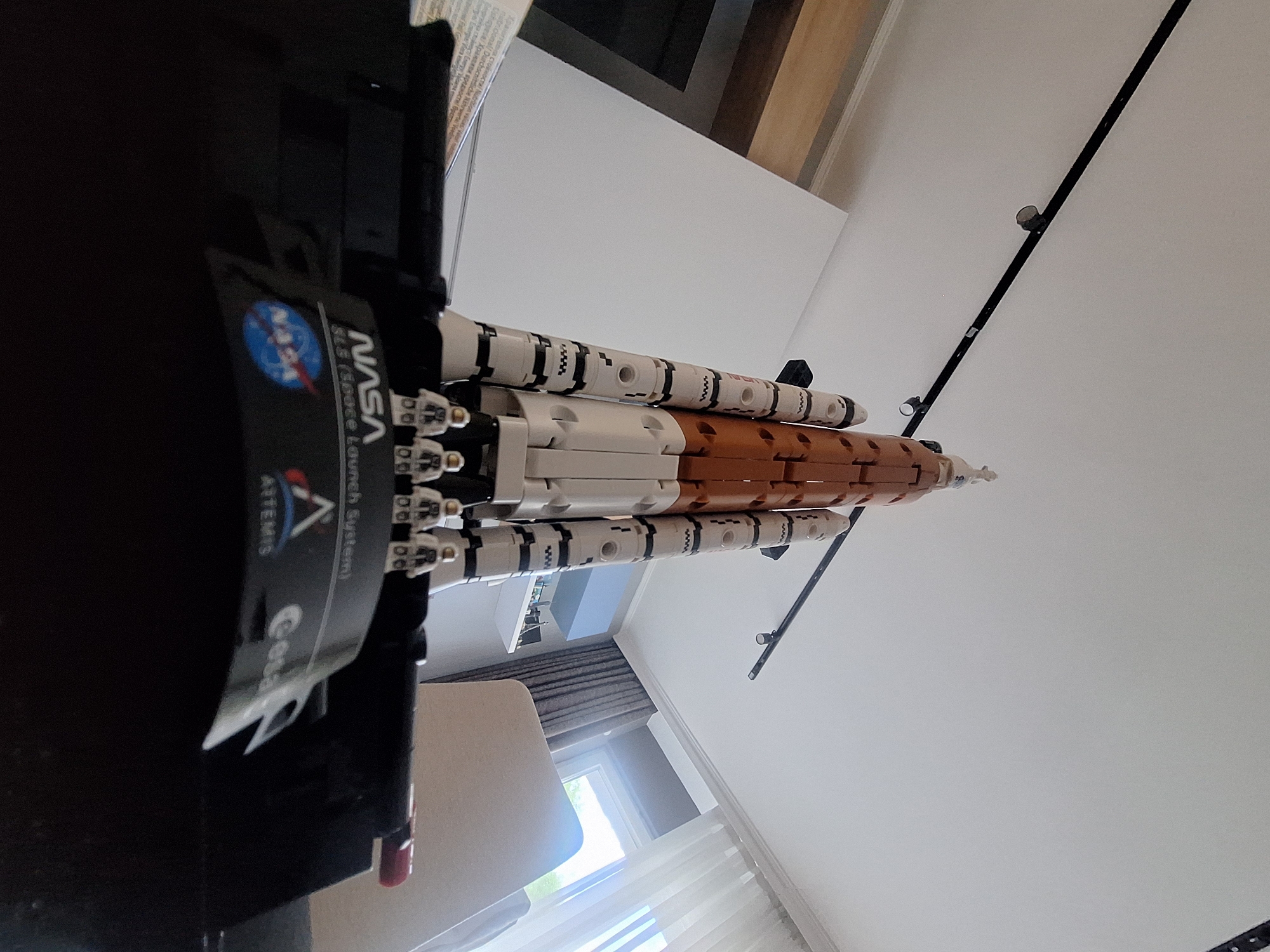 Here's a concise description of the image:
The image shows a Lego model of the NASA SLS (Space Launch System) rocket, displayed on a black stand with NASA and Artemis logos. The rocket itself is predominantly white and orange, with black and white patterned detailing on the boosters. The scene is indoors with a ceiling featuring a black track lighting system. A window with sheer white curtains is visible in the background. The image appears to be taken at an angle, looking up at the rocket. The surrounding environment includes a beige chair and a portion of the room's interior with light-colored walls. There is no specific information about the location being in Sofia, Bulgaria, or about any people present.