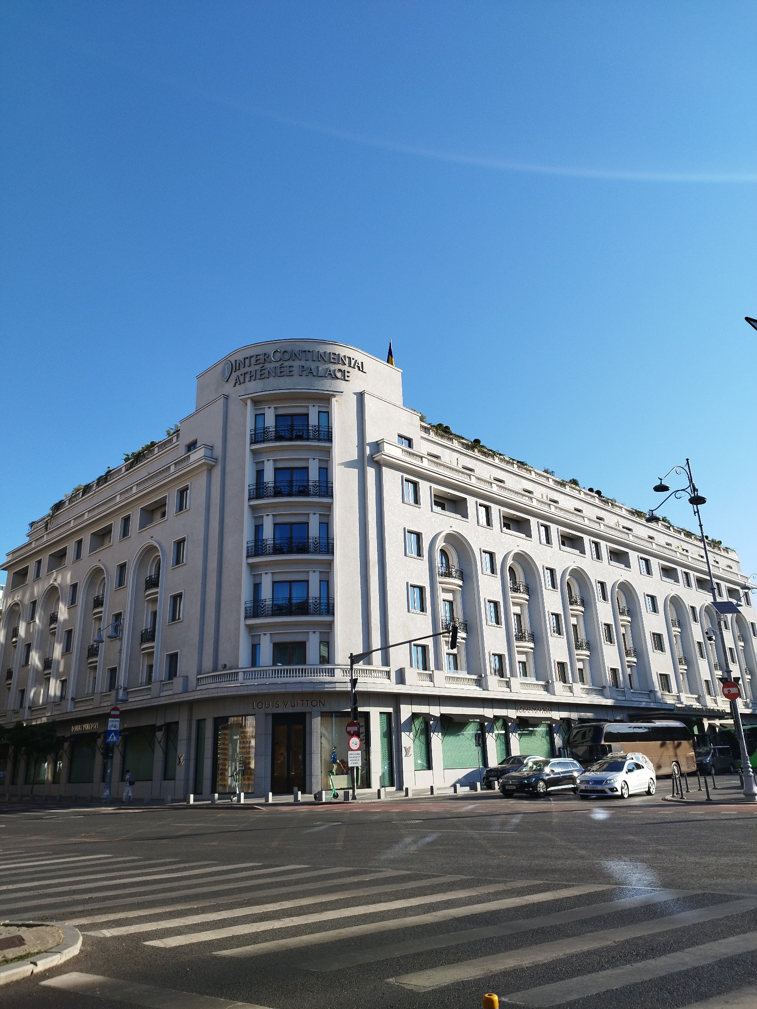 A wide-angle shot captures the InterContinental Athenée Palace hotel in Bucharest, Romania, on a bright, clear day. The grand, light-colored building dominates the frame, its multi-story facade featuring numerous arched windows with decorative balconies on the upper floors, and large display windows on the ground floor, one of which bears the "LOUIS VUITTON" signage. A black lamppost stands tall on the right side of the building, with a traffic light and a pedestrian crossing sign visible near the corner. Several cars are parked or driving on the street in front of the hotel, including a white sedan and a dark-colored SUV. A pedestrian can be seen walking on the sidewalk. The foreground shows a pedestrian crossing with white stripes on asphalt. The overall scene is one of urban elegance and activity under a clear blue sky.