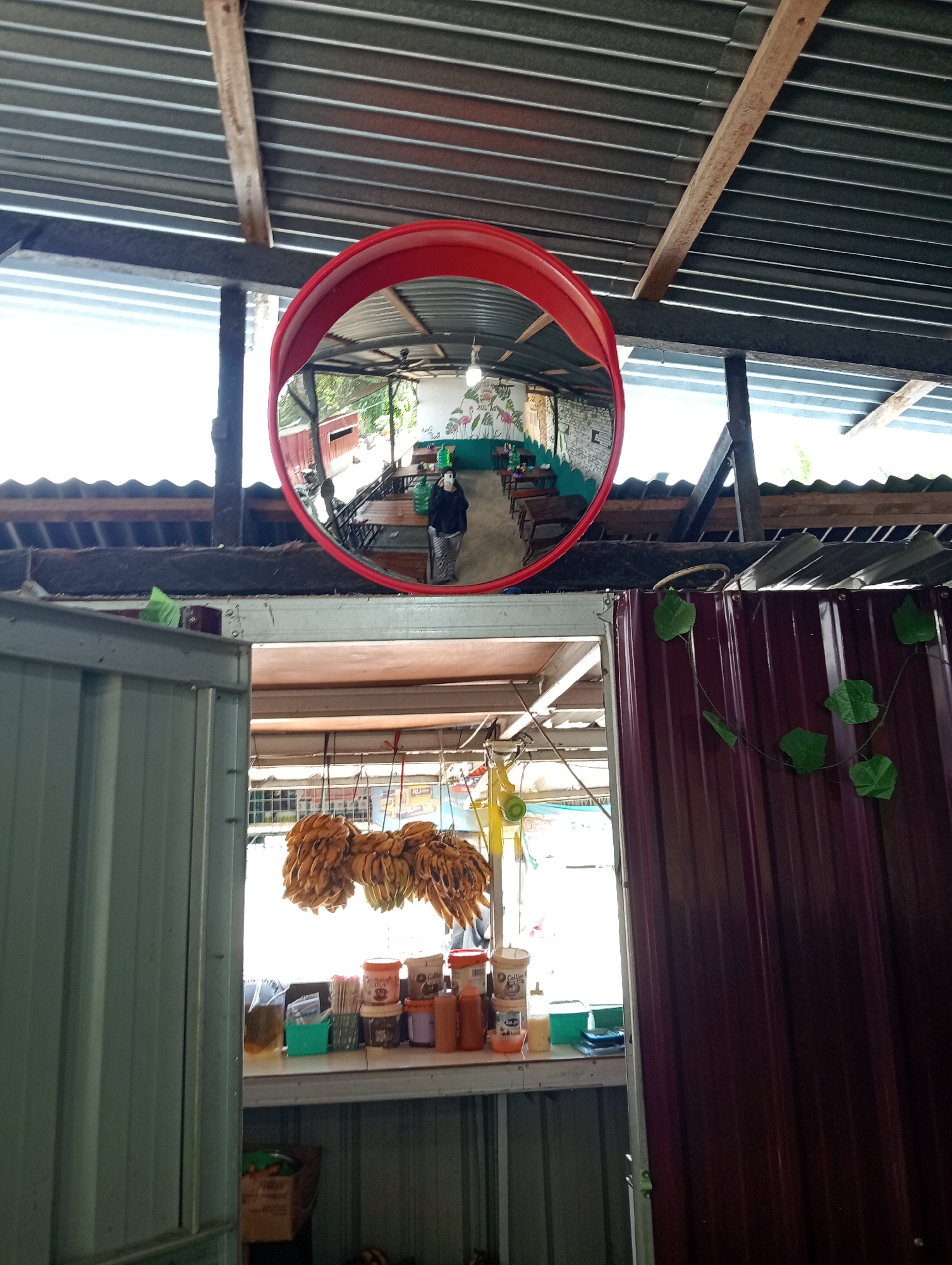 The image shows a wide-angle convex mirror mounted on a corrugated metal roof structure. The mirror reflects an interior scene of what appears to be an open-air food stall or small restaurant. The reflection shows several wooden tables and chairs, a ceiling fan, and a wall decorated with a mural of flamingos. A person wearing a black hoodie and striped pants is standing in the reflection, holding a phone, seemingly taking a selfie. In the foreground of the actual image, outside the mirror's reflection, is a stall displaying bunches of yellow bananas hanging from the ceiling and various containers on a shelf, including what appear to be jars of spices or condiments and cups. The overall environment suggests a casual, possibly tropical setting. The location is identified as Campago, Indonesia.