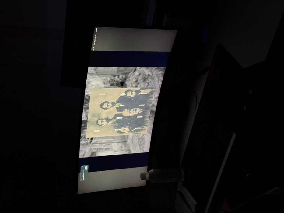 A dark room features a large monitor displaying an image of a black and white photo superimposed on a grayscale background. The photo shows four men in suits, with text boxes near each person. The monitor displays a typical interface with a menu bar at the top and a dock at the bottom, including a notification bubble. A water bottle and other items are visible near the base of the monitor, and a partially visible keyboard suggests a workspace setting.