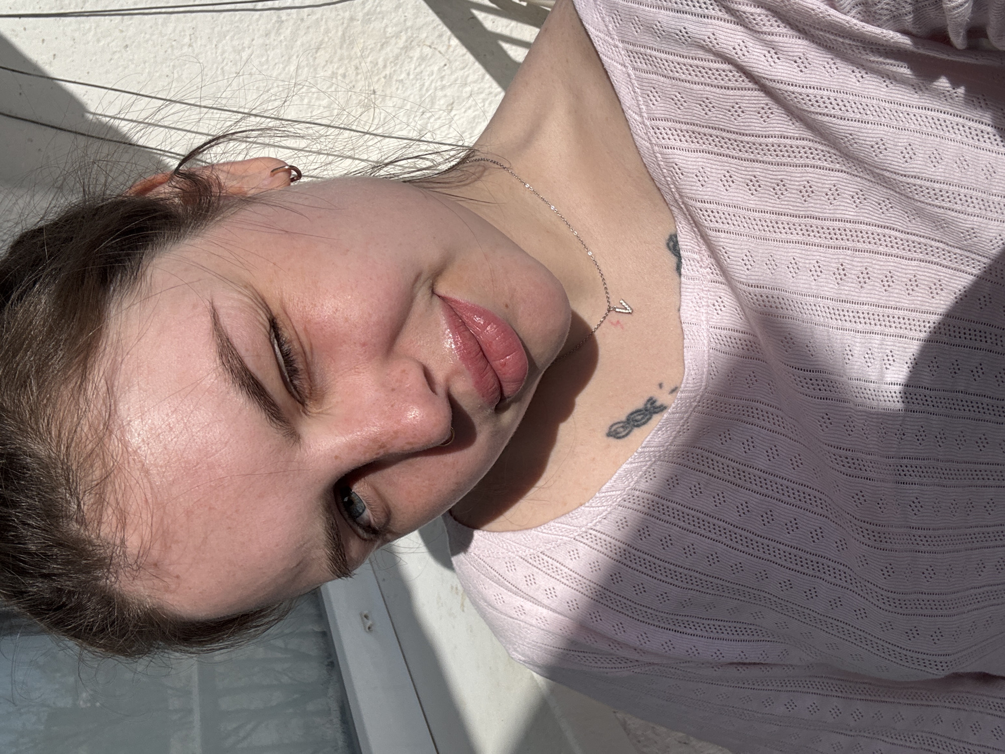 A close-up, high-angle shot of a fair-skinned woman. She wears a light pink, patterned, long-sleeved top. A silver necklace with a small "V" pendant is visible on her neck, along with two tattoos: one on her collarbone area and another on her upper chest. The woman has a nose piercing, dark eyebrows, and light blue eyes. The setting appears to be outdoors, with a white wall and a window frame partially visible. The lighting suggests bright sunlight. There is no evidence to suggest that this image was taken in Chornomorskoe, Ukraine.