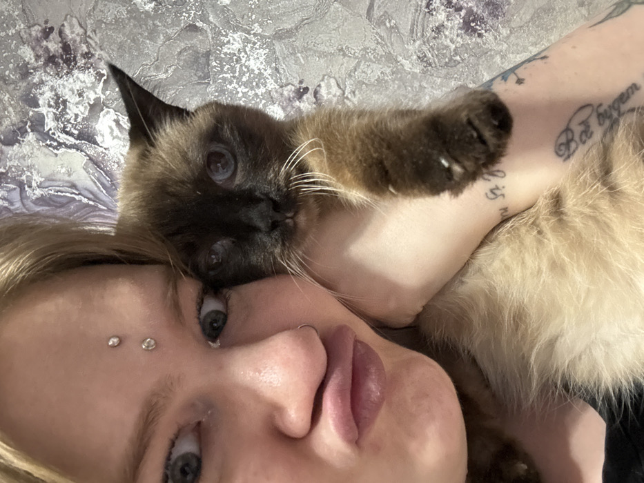 An indoor, close-up photograph depicts one individual holding a cat. The individual, appearing to be female, has light-colored hair, wide-set blue eyes, and a "duck face" expression with puckered lips. A silver-toned ring is present in the right nostril. Two clear, circular dermal piercings are visible vertically aligned on the forehead between the eyebrows. The individual's left arm is visible, revealing pale skin and multiple tattoos, including partially visible script near the wrist, a Cyrillic script tattoo further up the forearm (partially visible as "Всё будет"), and a faded design below it.
The cat is a Siamese or Siamese-mix, characterized by dark brown fur on its face, ears, and paws, and lighter tan fur on its body. It has blue eyes and prominent white whiskers, and is looking directly at the camera. Its right front paw rests on the individual's forearm.
The background is a wall covered with wallpaper that features an abstract, textured pattern in shades of grey, silver, and subtle purple, consistent with a domestic interior. The scene is located in Saratov, Russia.