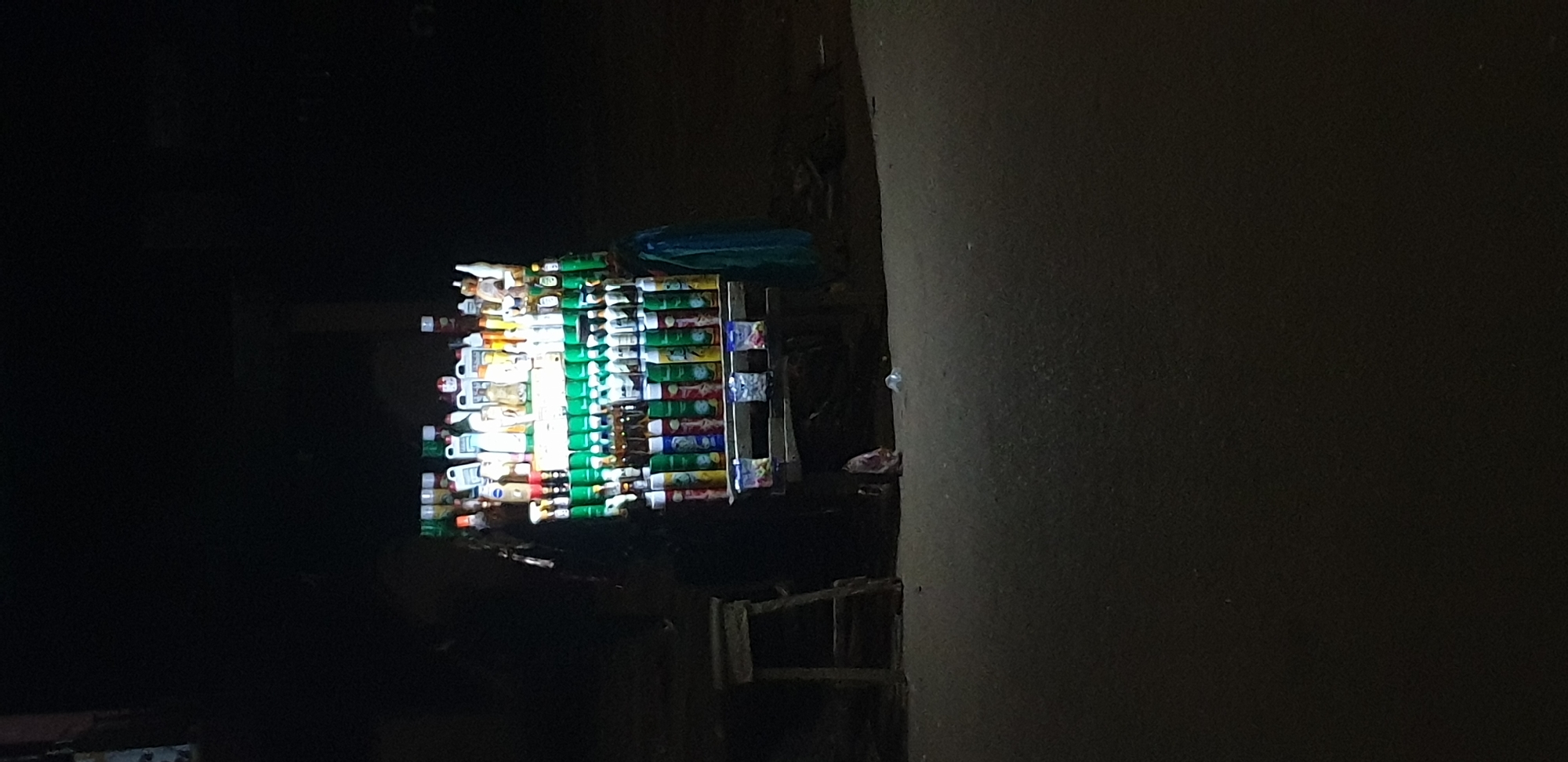 This image depicts a brightly illuminated vendor's stall at night on what appears to be a paved surface in Sabon Tasha, Nigeria. The stall, possibly a cart or fixed stand, is densely packed with various products arranged on multiple tiers.
The top tier displays numerous bottles of assorted shapes, sizes, and colors, suggestive of toiletries, lotions, or household liquids. Below this, a prominent tier features two rows: an upper row dominated by uniform green aerosol cans, and a lower row containing a mix of brown bottles, white bottles, and more aerosol cans. The lowest visible tier showcases multiple multicolored aerosol cans (green, red, blue, yellow) and, at the base of the stall, several small, colorful packaged items, possibly sachets, are visible.
A dark, indistinct human figure stands or sits to the left of the stall, partially obscured by shadow, likely attending to the stand. To the immediate left of the stall, a rudimentary wooden stool with a flat top and splayed legs is positioned on the paved ground. A light-colored plastic bottle or object lies on the ground near the stool, and a small, reddish-pink object, possibly litter, is visible closer to the stall. A blue or green fabric hangs from the right side of the stall, reaching the ground. The immediate foreground shows a dark, textured paved surface, while the background and areas surrounding the stall are enveloped in deep shadow, rendering details indistinguishable. The primary light source emanates directly from the stall, highlighting its contents against the pervasive darkness of the night.
