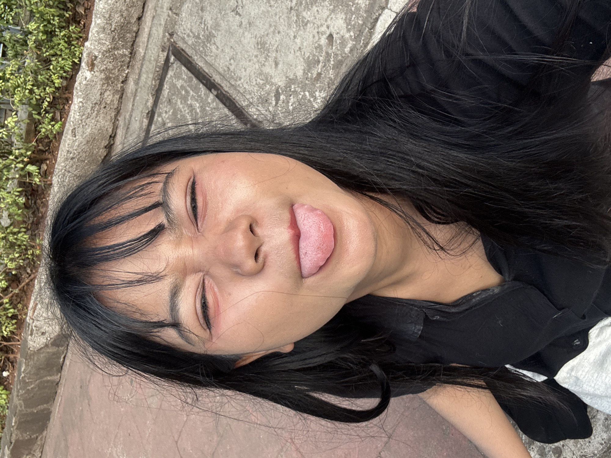 A young woman with black hair and bangs is pictured from the chest up, outdoors on a paved area with red brick and concrete flooring. She is wearing a black collared shirt. Her eyes are squinted shut, and her tongue is sticking out. The background shows a stone border and some green foliage, consistent with a public park or outdoor seating area in West Jakarta, Indonesia.