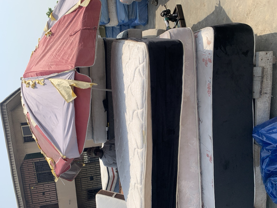 The scene depicts an outdoor commercial display in Alaiyabiagba, Nigeria, under bright direct sunlight. In the foreground and midground, a vertical stack of four mattresses dominates the frame. The uppermost mattress features a white quilted top and black side panels with white piping. Below it are two mattresses exhibiting light-colored, possibly beige or pink, floral-patterned fabric. The bottom mattress, resting on visible wooden pallets, also has a white quilted top with black side panels and white piping.
Providing partial shade over the mattress stack is a large, weathered market umbrella. Its fabric is faded red and light purple/grey, with numerous torn yellow strips of material.
Behind the umbrella and the central mattress stack, a male individual is partially visible. He wears a light-colored short-sleeved collared shirt with a dark logo (partially obscured, possibly "JUVAH") and a dark beanie hat with visible text (partially obscured, possibly "BOSS"). He appears to be looking forward.
To the right of the main mattress stack, a small, dark mechanical device, resembling a hand-cranked sewing or upholstery tool, rests on a black tray on the ground. In the background, a multi-story building with a light-colored facade and dark-framed windows with security bars is visible. Further to the right in the background, additional stacks of mattresses, some wrapped in blue plastic, are present. A crumpled blue tarp or plastic sheet lies on the ground in the lower-left foreground. The ground surface is light-colored, consistent with concrete or compacted earth.