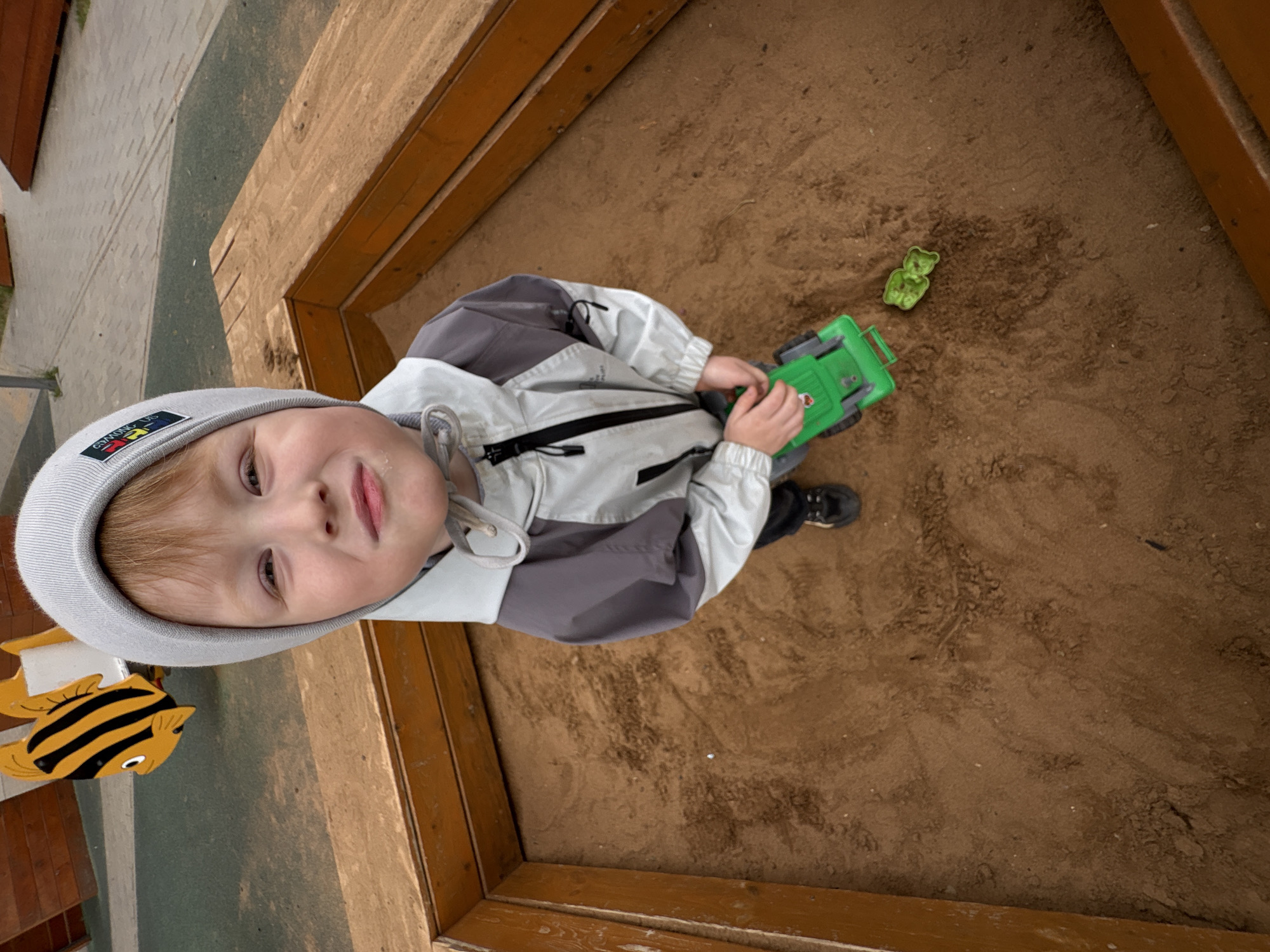 This image captures a young child in a sandbox on a playground in Izhevsk, Russia. The child, a boy with light reddish-brown hair, wears a light gray beanie with an "AMONG US" patch and a two-tone jacket, light gray on top and darker gray on the bottom, featuring a black zipper. He stands in the center of the wooden-framed sandbox, holding a green toy truck in his left hand. The boy looks directly at the camera with a slight smile, and his face shows traces of sand. The sandbox is filled with light brown sand, exhibiting various textures and disturbed areas. A small, green, leaf-shaped object lies on the sand to the child's right. In the background, beyond the sandbox, a yellow and black striped fish-shaped spring rider is visible on a rubberized playground surface, which transitions into patterned paving. The overall setting is an outdoor recreational area.