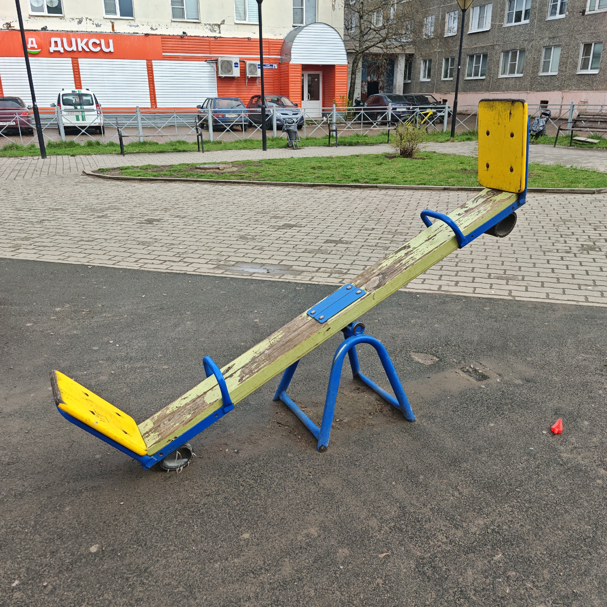 An outdoor urban scene is depicted, focusing on a weathered seesaw in the foreground, likely situated in a playground or courtyard. The seesaw features bright yellow seats and handles, a light green wooden plank that is chipped and worn, and a blue metal base. One end of the seesaw is elevated, while the other is closer to the ground. The immediate ground surface is dark grey asphalt or a similar rubberized material, showing patches of wear and some debris. A small, red plastic object lies on the ground to the right of the seesaw.
Beyond the seesaw, the ground changes to light grey paver stones, which border a narrow strip of green grass. A metal fence separates this area from a street and buildings in the background. On the left side, an orange and white commercial building is visible, featuring the Russian word "ДИКСИ" (DIKSI) in red letters. Several cars are parked behind the fence in front of this building; one white car displays the license plate "702BY578", and a dark car has "H 122...". An air conditioning unit is mounted on the building's facade. To the right, another entrance with a rounded canopy is visible, followed by a multi-story grey residential building with numerous windows. More cars are parked in front of this building.
There are no people present, and no specific activity is taking place. The scene appears to be set during daytime on an overcast day, indicated by the diffused light. The visible text on the store, "ДИКСИ", is a prominent Russian supermarket chain, consistent with the given location context of Rostov, Russia.
