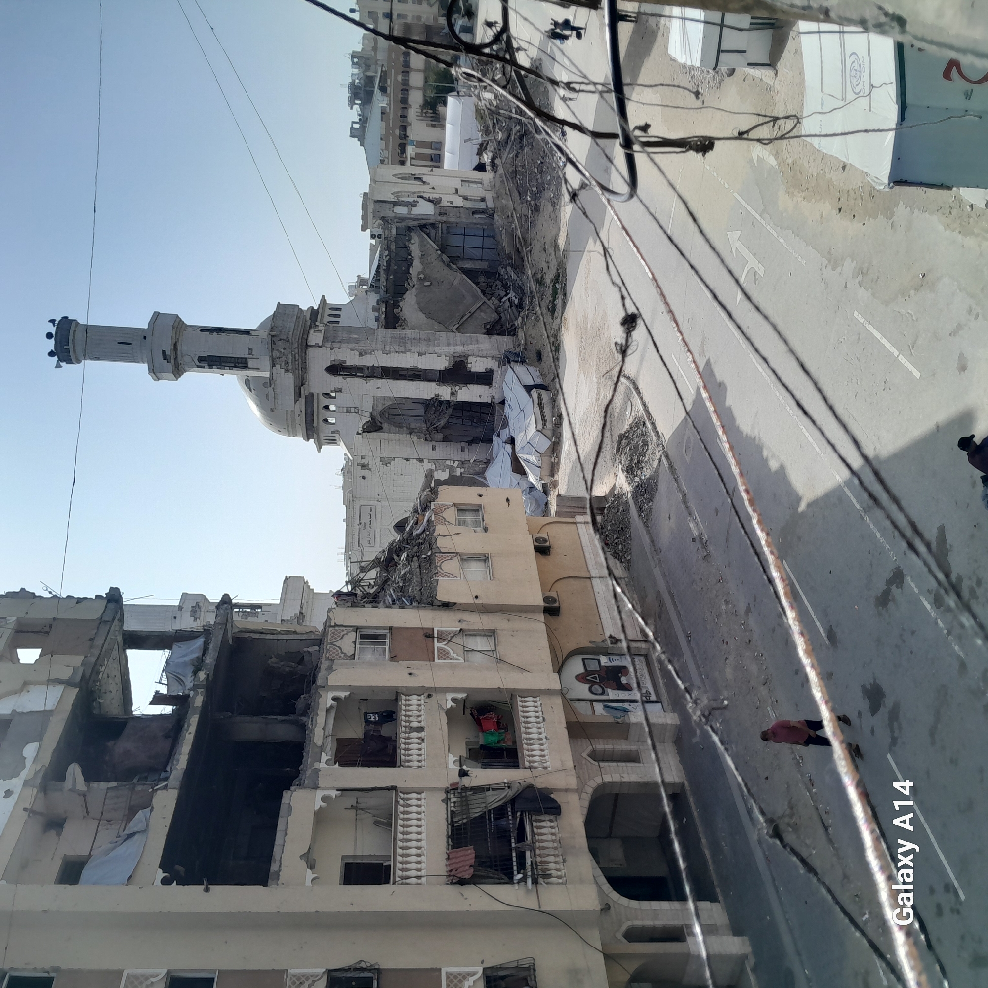 The image, oriented vertically, depicts a street scene of an urban area heavily affected by damage, likely from conflict or a disaster. In the background, a prominent white mosque with a tall minaret and a large dome stands, appearing partially damaged or in a state of disrepair, with visible cracks and missing sections.
Along the street, several multi-story residential buildings show extensive structural damage. Many have large sections of their facades and interiors blown out, revealing rubble, exposed rooms, and debris. White tarpaulins are draped over some of the damaged sections of buildings and over piles of rubble on the ground, possibly for protection or to cover displaced materials.
The paved street in the midground features white lane markings, including a white arrow indicating a turn. The ground adjacent to the street is covered in dirt and debris. Numerous utility lines or electrical wires crisscross overhead, casting distinct shadows on the street below.
One person is visible in the lower right portion of the image, walking away from the viewer on the street, wearing a dark top and lighter trousers. Another darker figure can be seen further down the street.
The time of day appears to be bright and sunny, indicated by the clear sky and sharp shadows cast by buildings and overhead wires. The overall atmosphere conveys a sense of devastation and the aftermath of a significant event.
Visible text includes a "Galaxy A14" watermark in the bottom right corner of the original image, indicating the device used to capture it. On one of the white tarpaulins covering debris, the acronym "IOM.OIM" is clearly visible, referencing the International Organization for Migration. Another tarp has a partial "2" on it. Some unreadable Arabic script is faintly visible on the mosque structure.