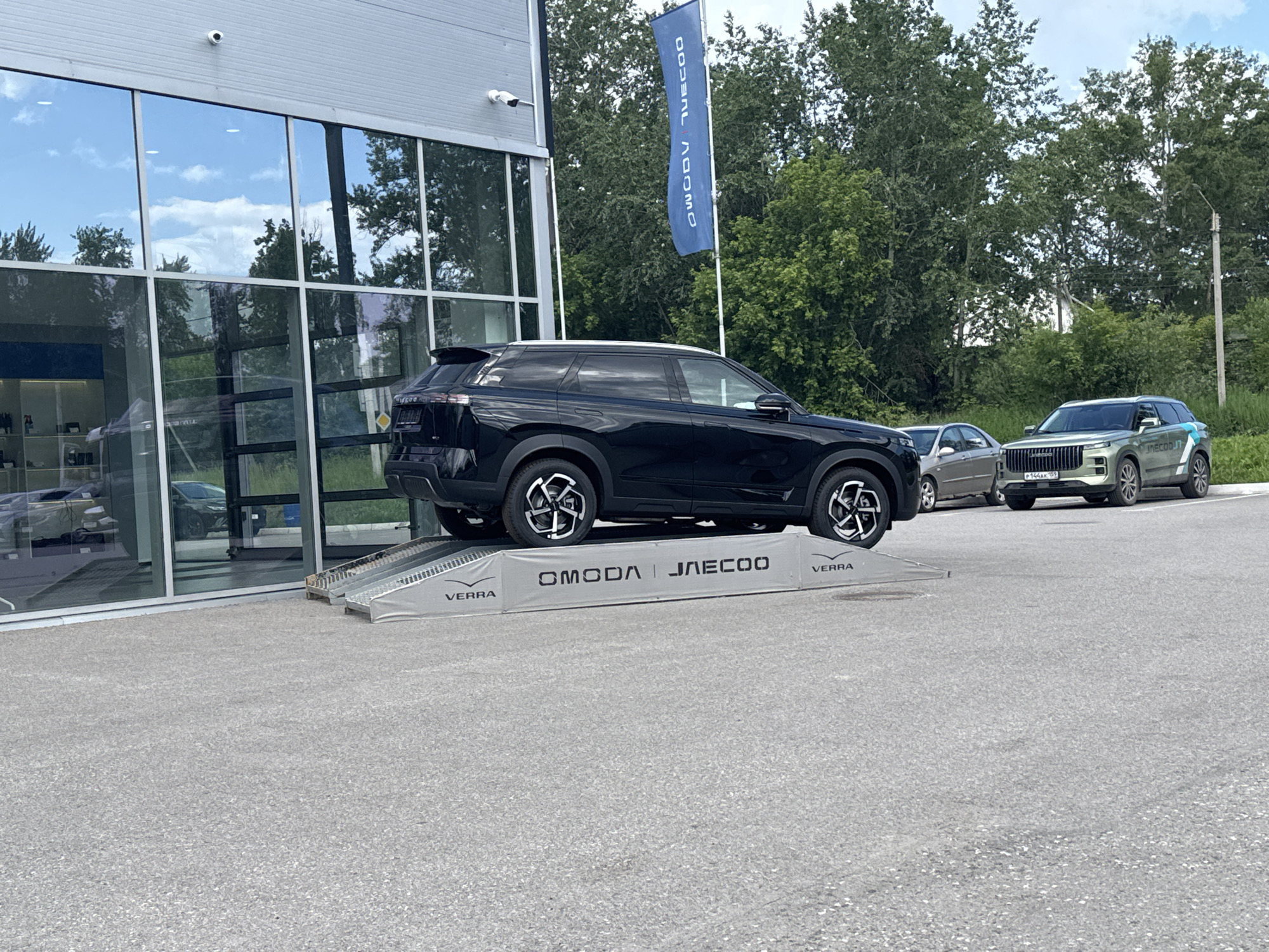 In the city of Berezniki, Russia, a black OMODA JAECOO SUV is parked on a grey ramp in front of a modern building with large glass windows. The ramp has "VERRA" and "OMODA JAECOO" branding. To the right of the black SUV, a lighter colored SUV with the same branding is parked on the asphalt. In the background, trees and a blue flag with white text are visible. The scene appears to be in a car dealership or service area.