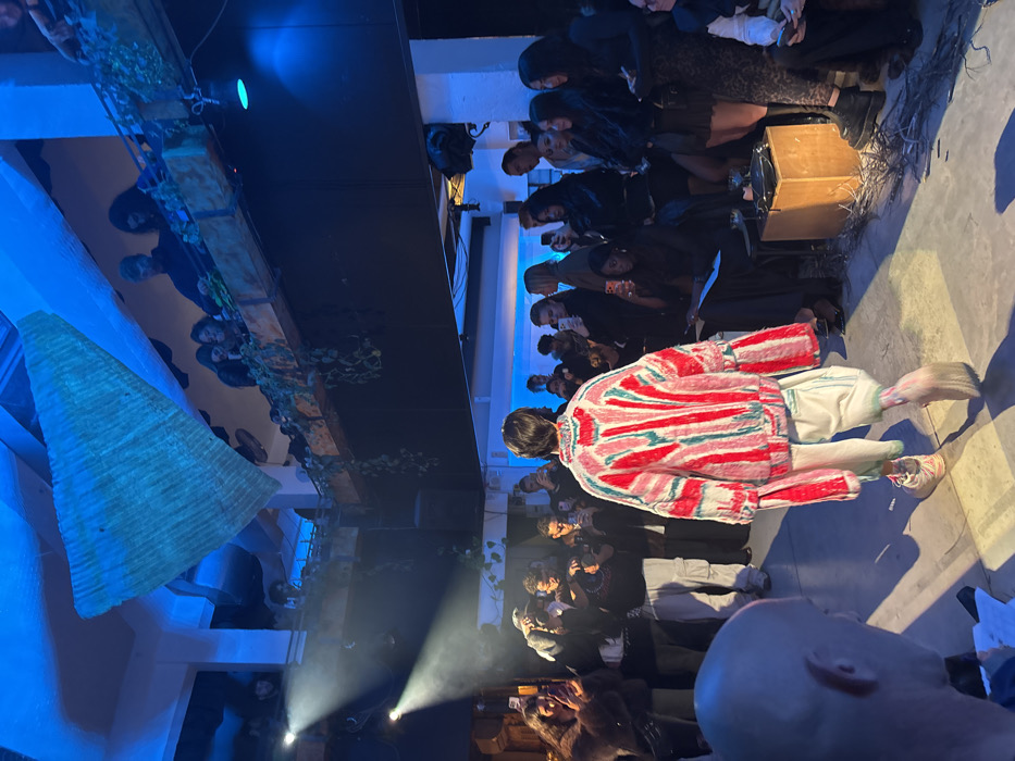 A fashion show with a model wearing a red and white striped jacket and white shorts. The model is walking down a runway, surrounded by an audience holding up phones to capture the moment. The room has a blue hue, with spotlights shining on the model. A large, triangular, woven structure hangs from the ceiling, adding to the artistic ambiance.