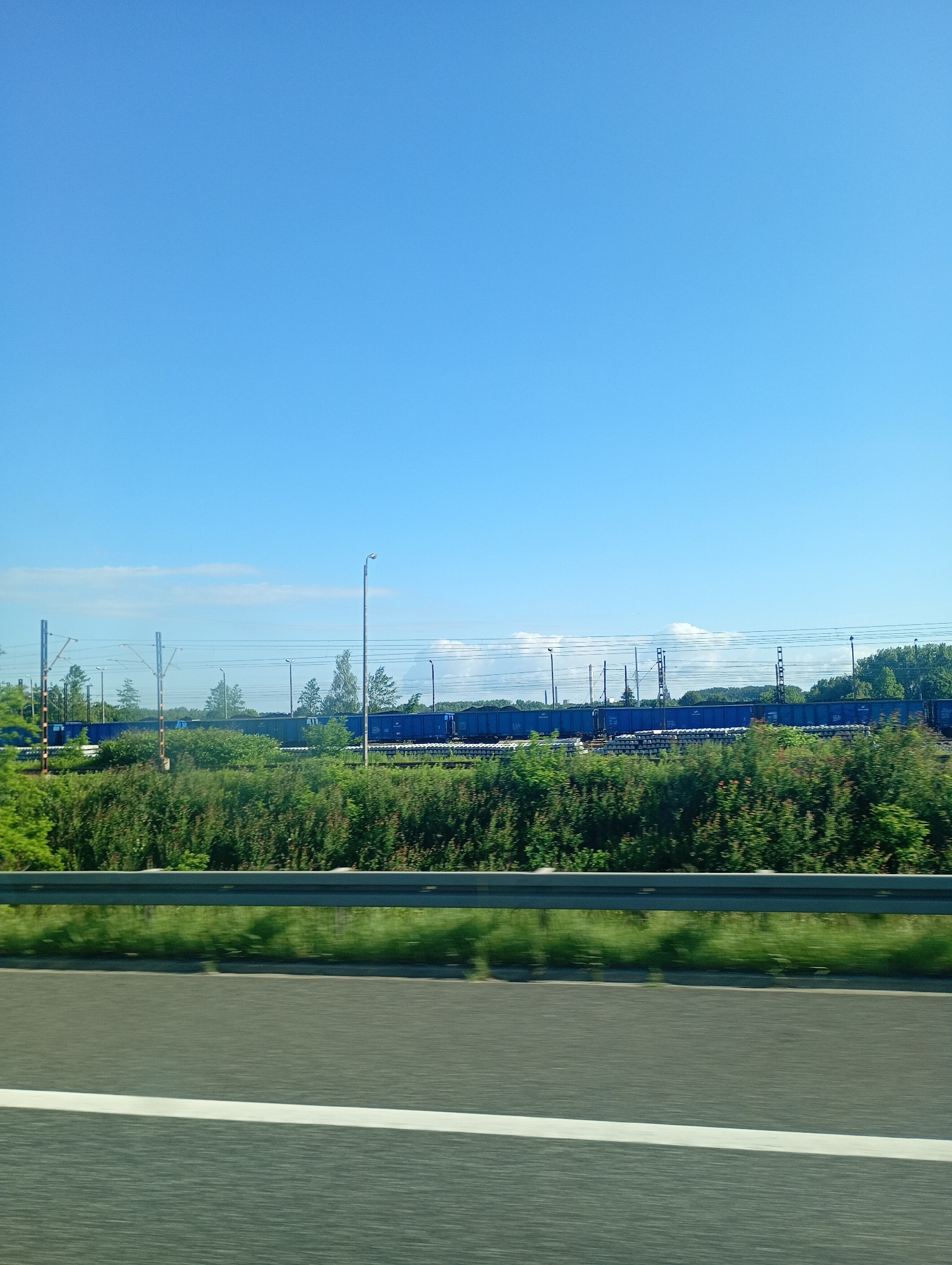 The image shows a view from a vehicle traveling on a highway in Ruda Śląska, Poland. In the background, a long train composed of blue freight cars is visible on a railway track. The train is stationary and appears to be loaded with materials, some of which are covered. The railway line is lined with lush green bushes and trees. Overhead power lines for the train are also present, supported by metal poles. The sky is clear and blue with a few wispy clouds. In the foreground, the highway barrier and road surface are visible, indicating movement. The overall scene depicts an industrial or transportation hub within a natural landscape.