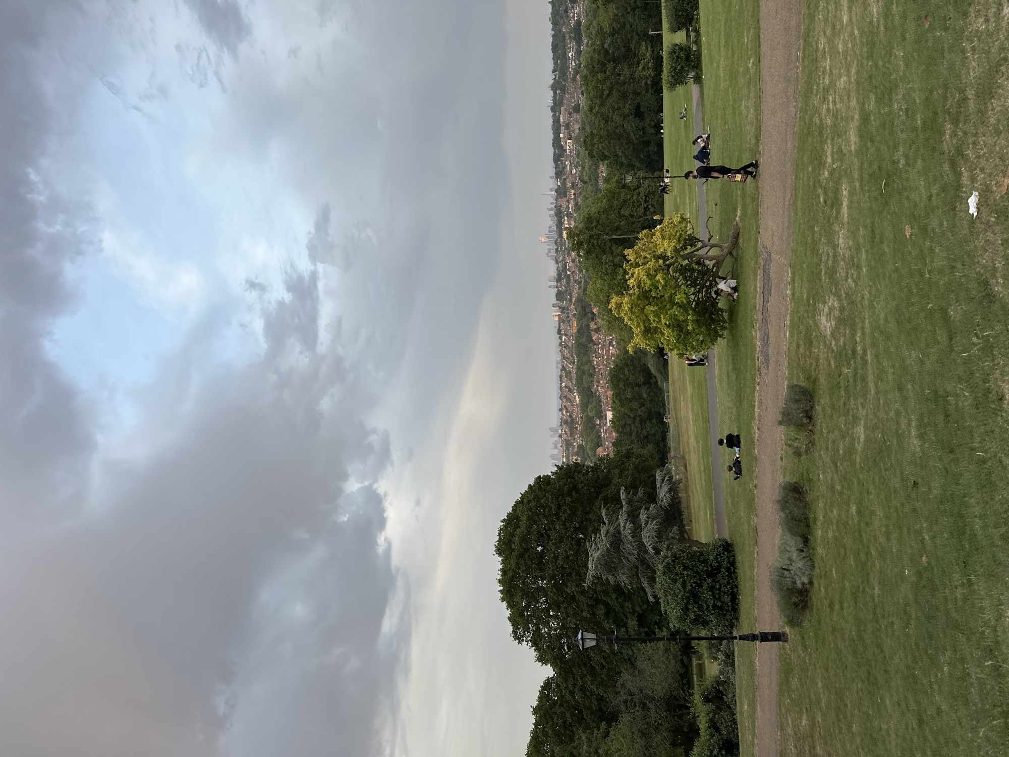 The image depicts a park on a cloudy day, possibly in the city of London due to the skyline visible in the background. The park features green grass, a gravel path, trees, and lampposts. There are several people in the park, some sitting and relaxing on the grass and others walking along the path. A large, distinctive tree with yellow-green foliage is prominent in the foreground. The sky is dominated by dark and light gray clouds, suggesting potentially inclement weather. In the distance, a city skyline is visible amidst the trees. There is some litter on the grass in the foreground.