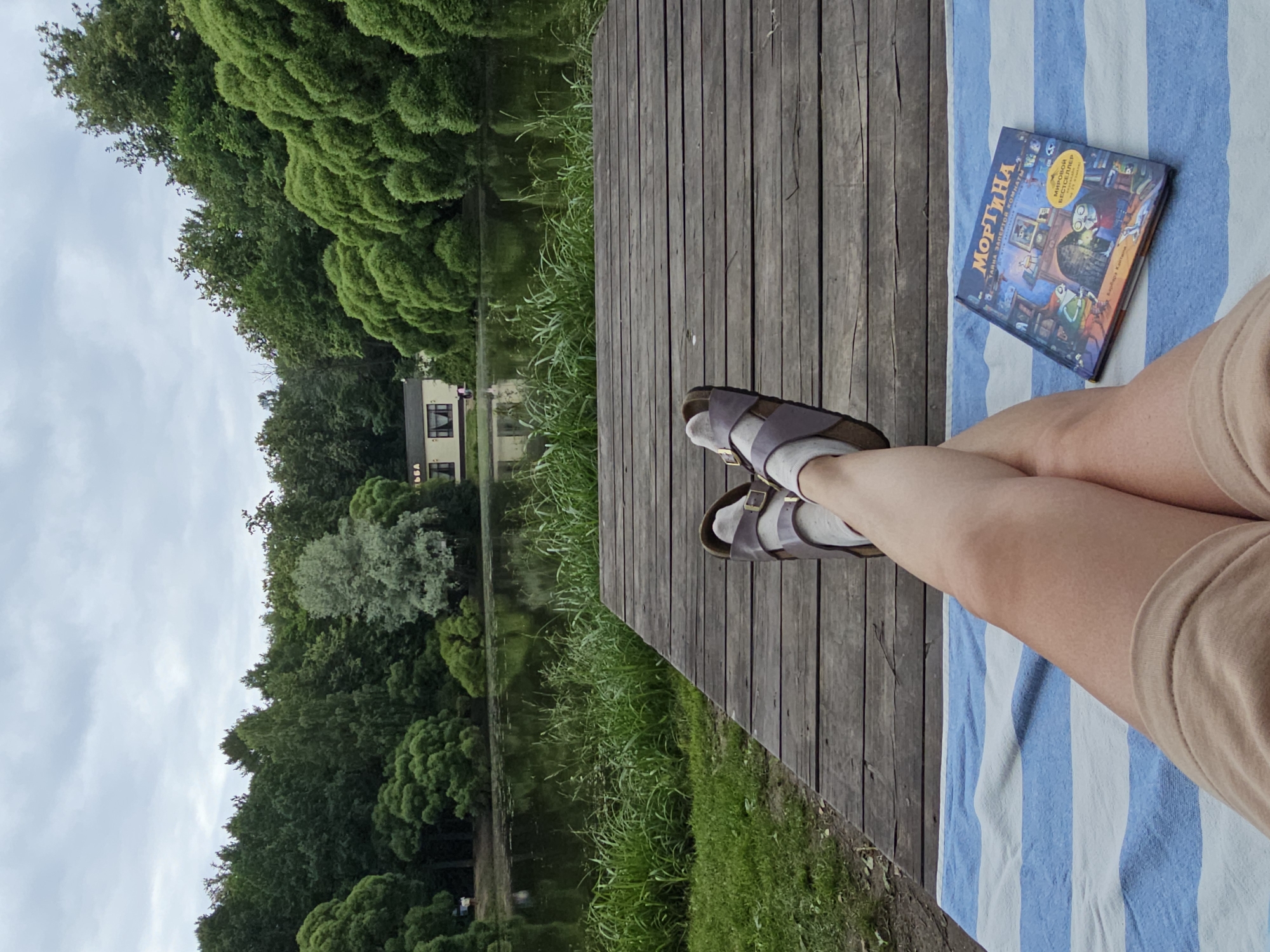 A person's legs, clad in beige shorts and white socks with brown Birkenstock sandals, are crossed as they relax on a blue and white striped blanket on a wooden deck. To the right, a book with a colorful cover depicting characters and a fireplace is placed on the blanket. The deck overlooks a body of water, likely a lake or pond, surrounded by lush green trees and foliage. In the background, a small building with a light-colored facade and a dark roof is visible near the water's edge. The sky is overcast with clouds. The scene appears to be a peaceful outdoor setting, possibly a park or recreational area, and the presence of water and abundant greenery suggests a natural environment within or near Moscow, Russia.