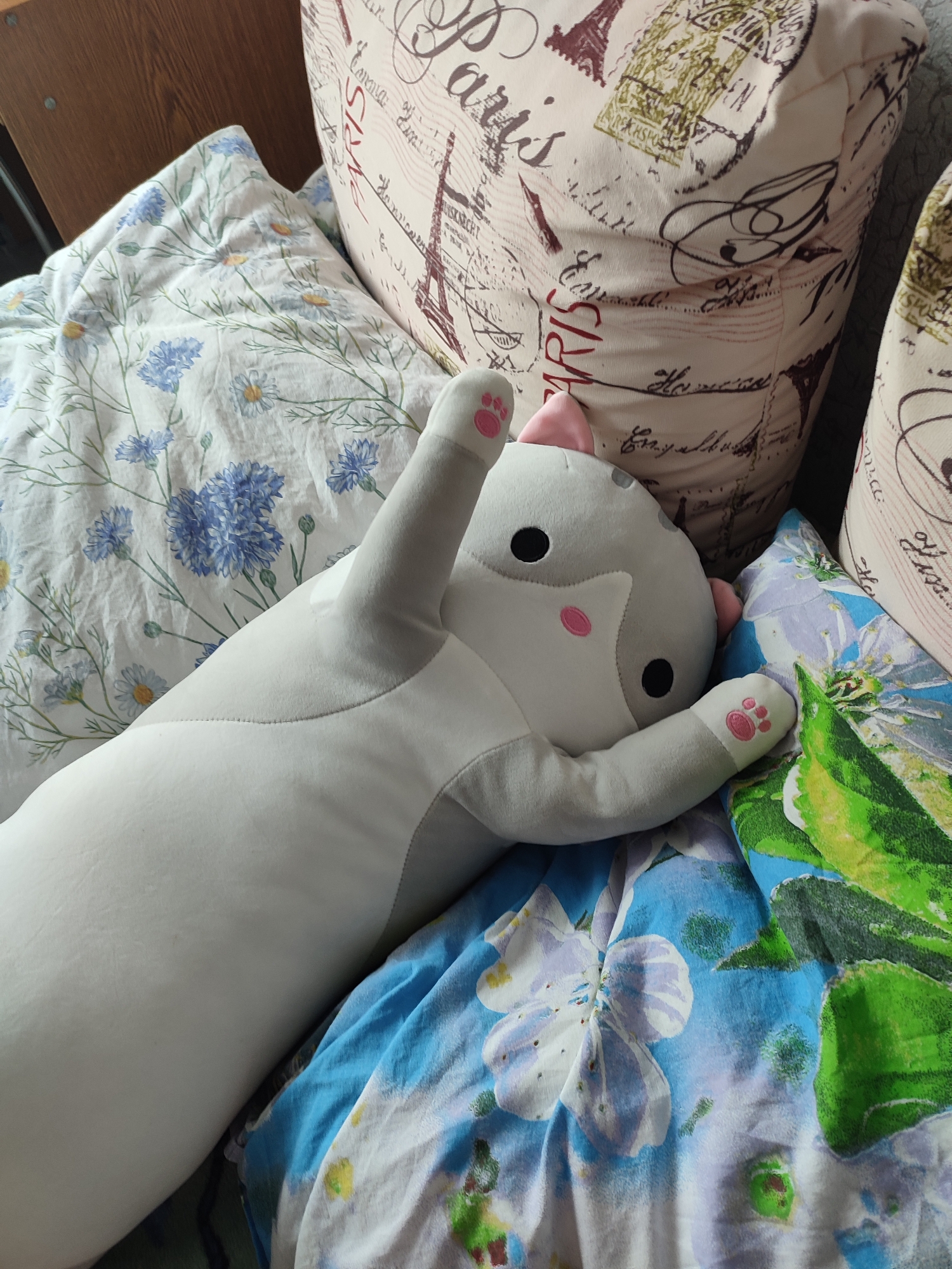 A plush cat toy lies on a bed, partially obscuring a floral patterned comforter. The cat toy is gray and white with pink paws and ears, and its eyes are black. The comforter features a pattern of blue flowers and white daisies. In the background, there are pillows with a French postcard design, depicting the Eiffel Tower and script writing. The scene appears to be indoors, likely a bedroom. There is no indication of the location being in Narowlya, Belarus.